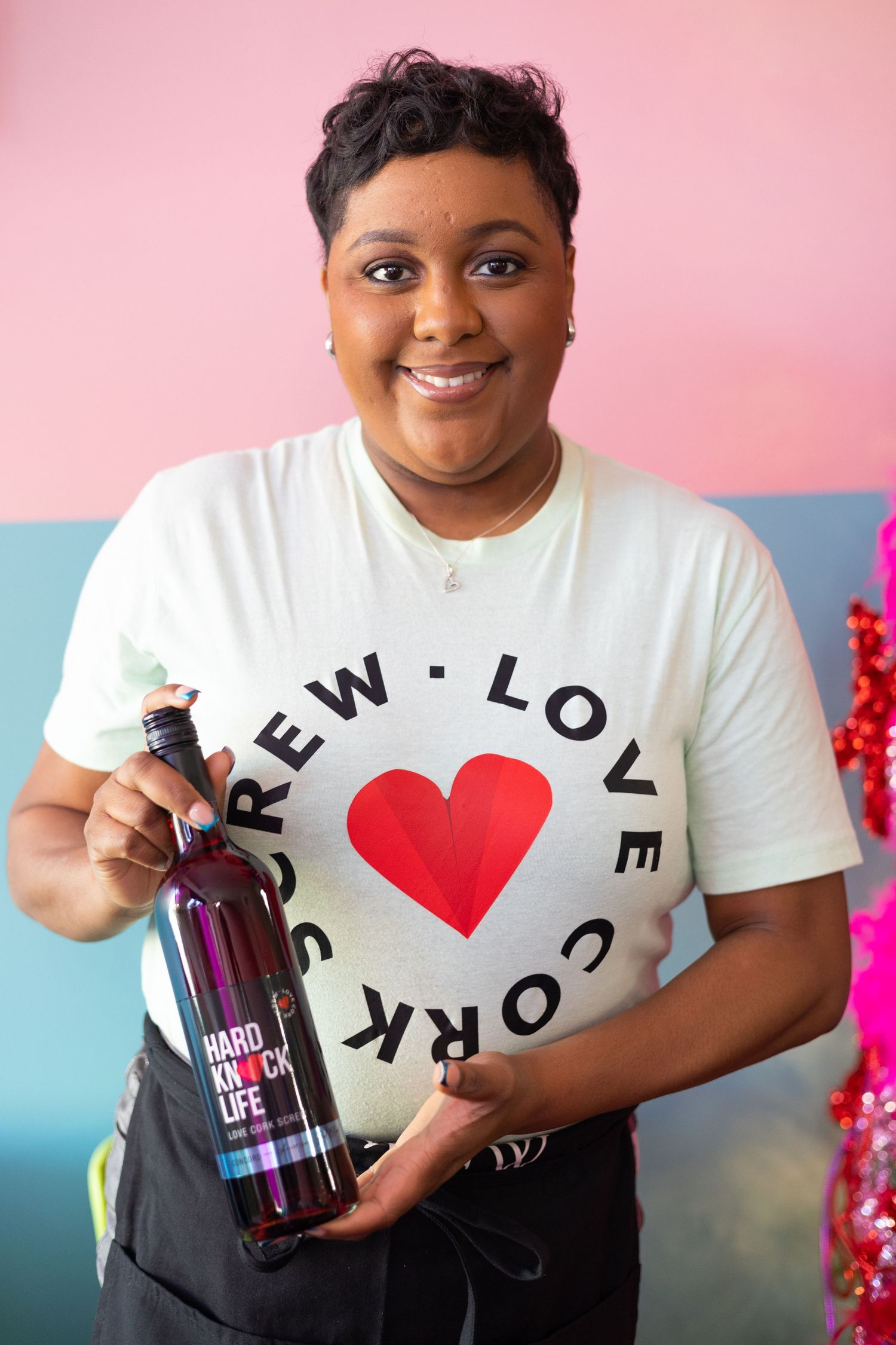 Person in white t-shirt with heart logo holding dark bottle against pink and blue background.