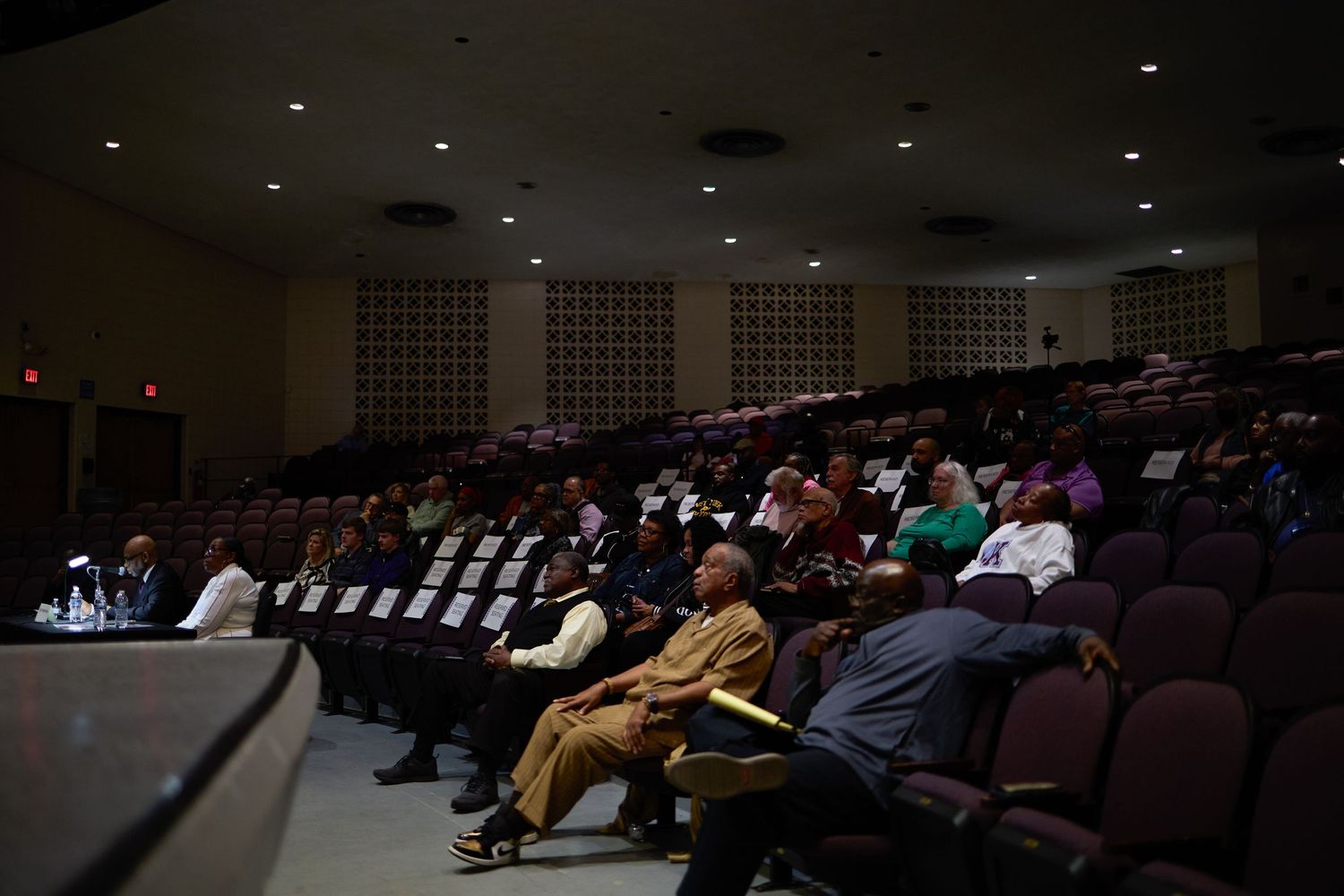 People seated in a dimly lit auditorium with rows of brown chairs during a presentation or event.