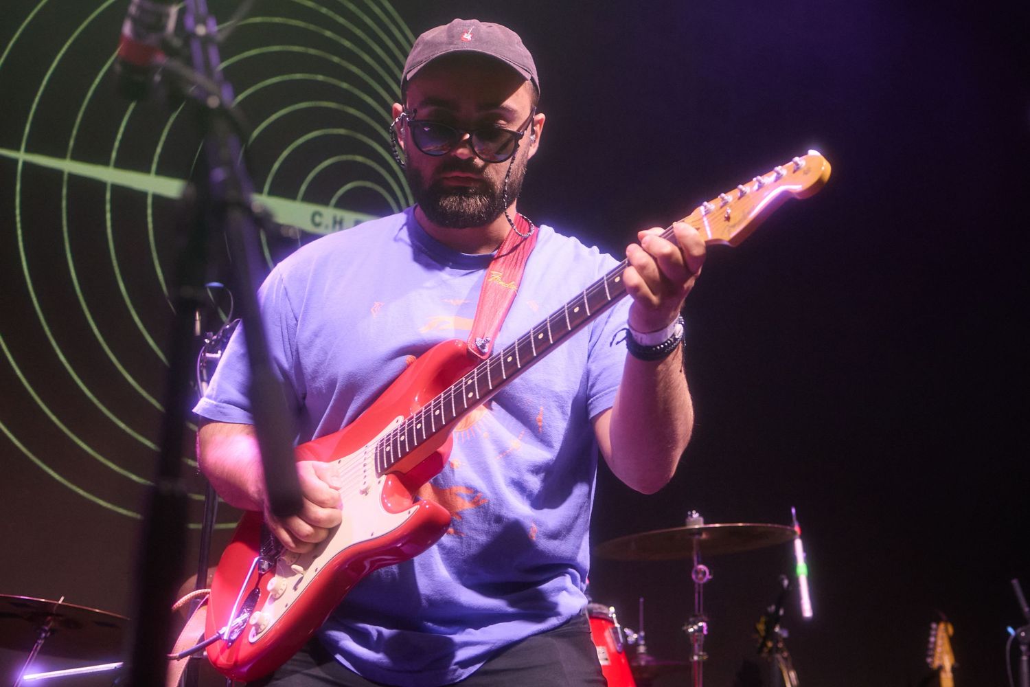 Guitarist in white t-shirt performs on stage with red electric guitar under dramatic lighting.