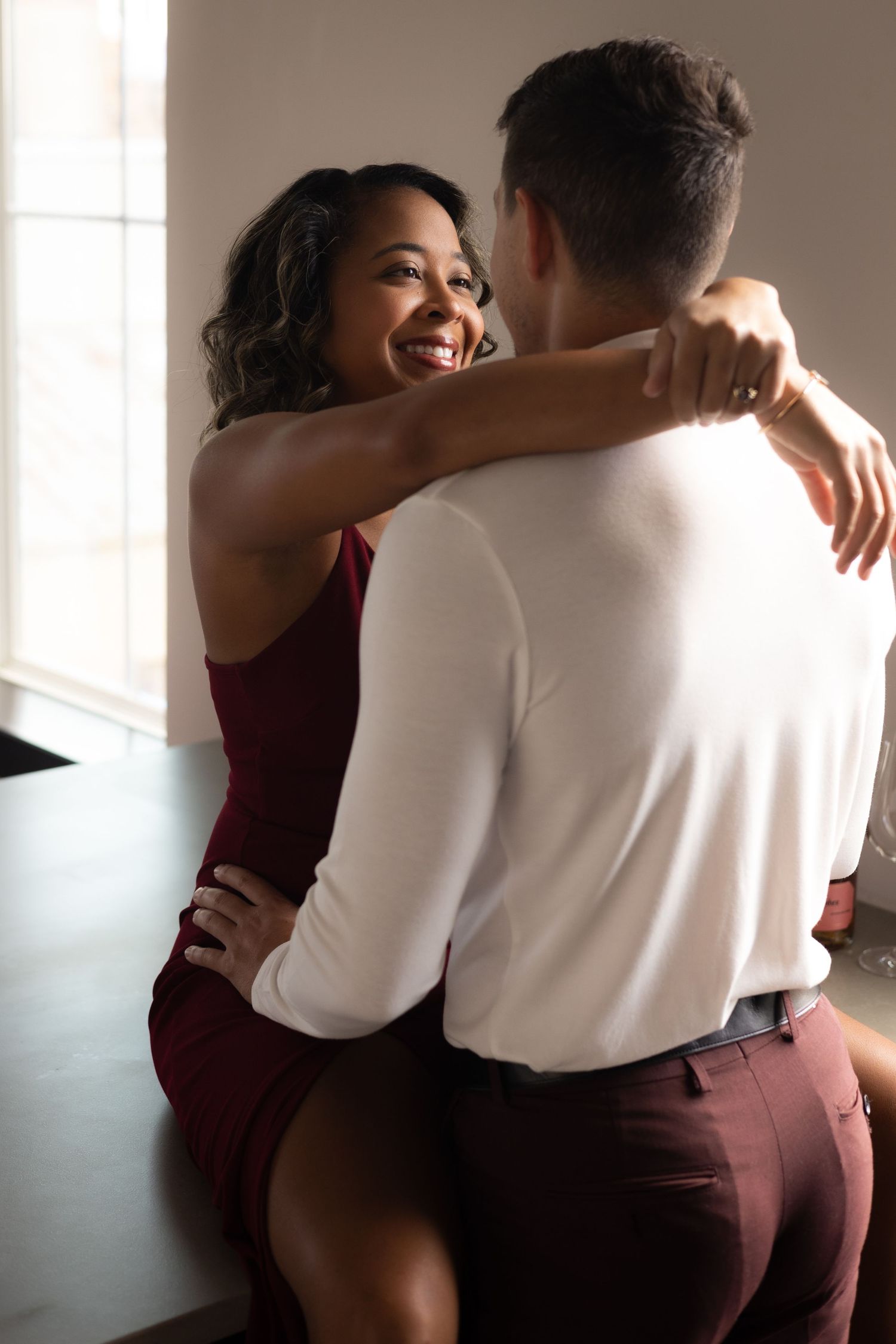 A couple shares an intimate moment together in burgundy and white outfits near a window with soft lighting.