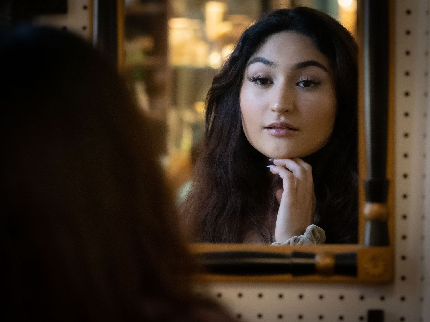 Portrait in warm lighting shows someone with dark hair and makeup against a blurred background mirror interior setting.
