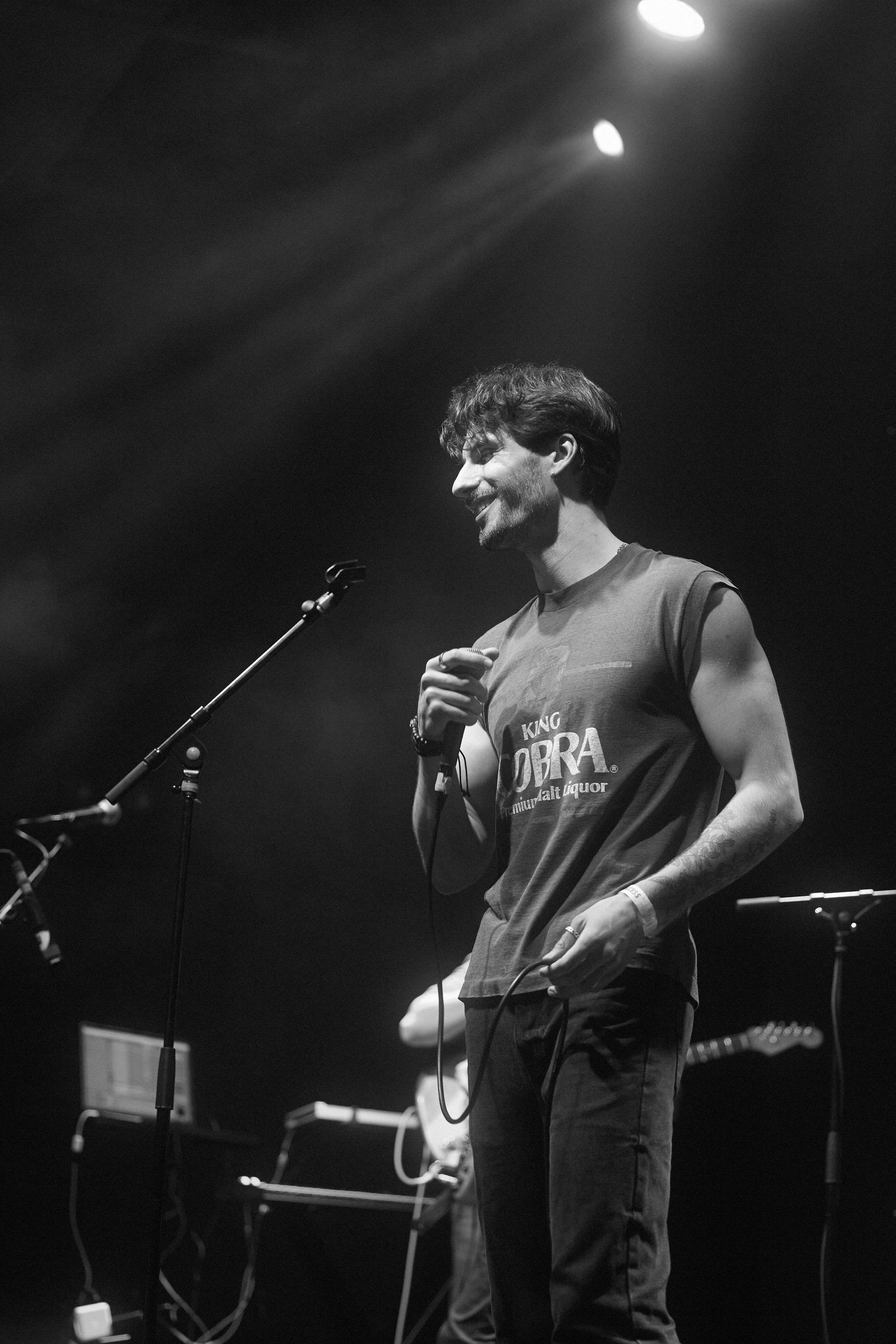 A performer in a sleeveless shirt sings into a microphone on stage with dramatic spotlights illuminating the scene.