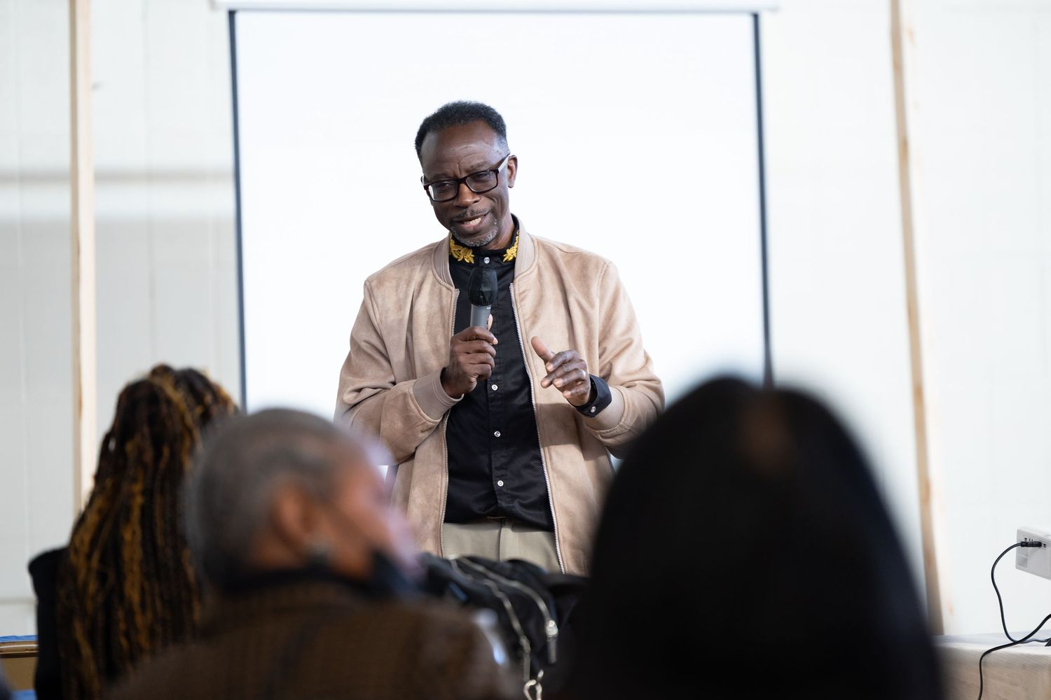A speaker wearing a beige jacket and glasses presents to an audience in a bright indoor setting.