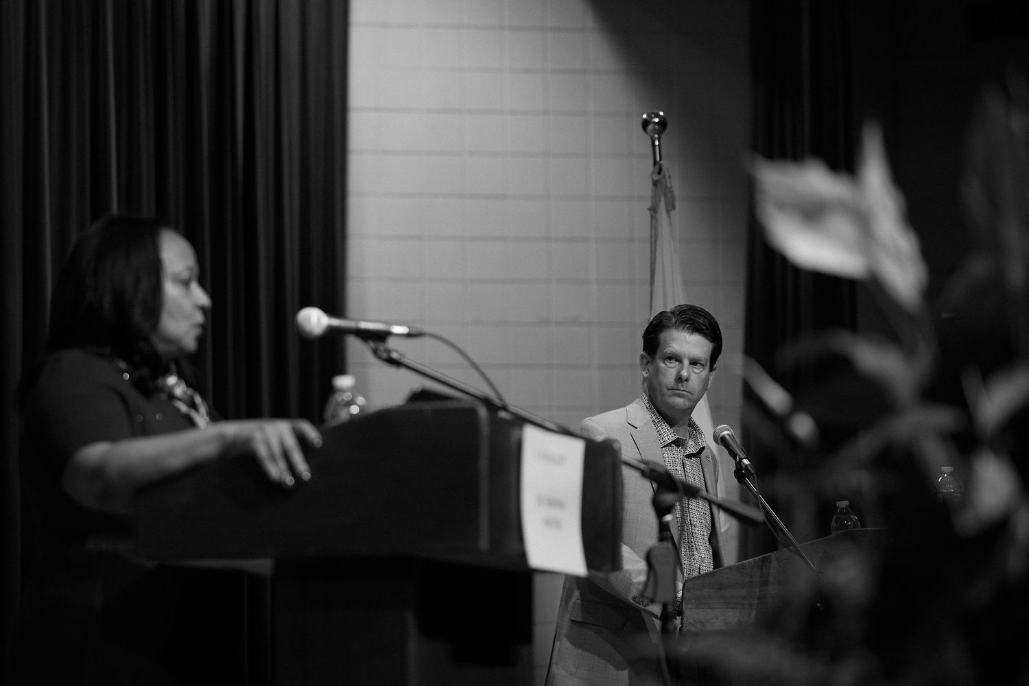 Black and white photo of two speakers at podiums during a public event with flags visible in the background.
