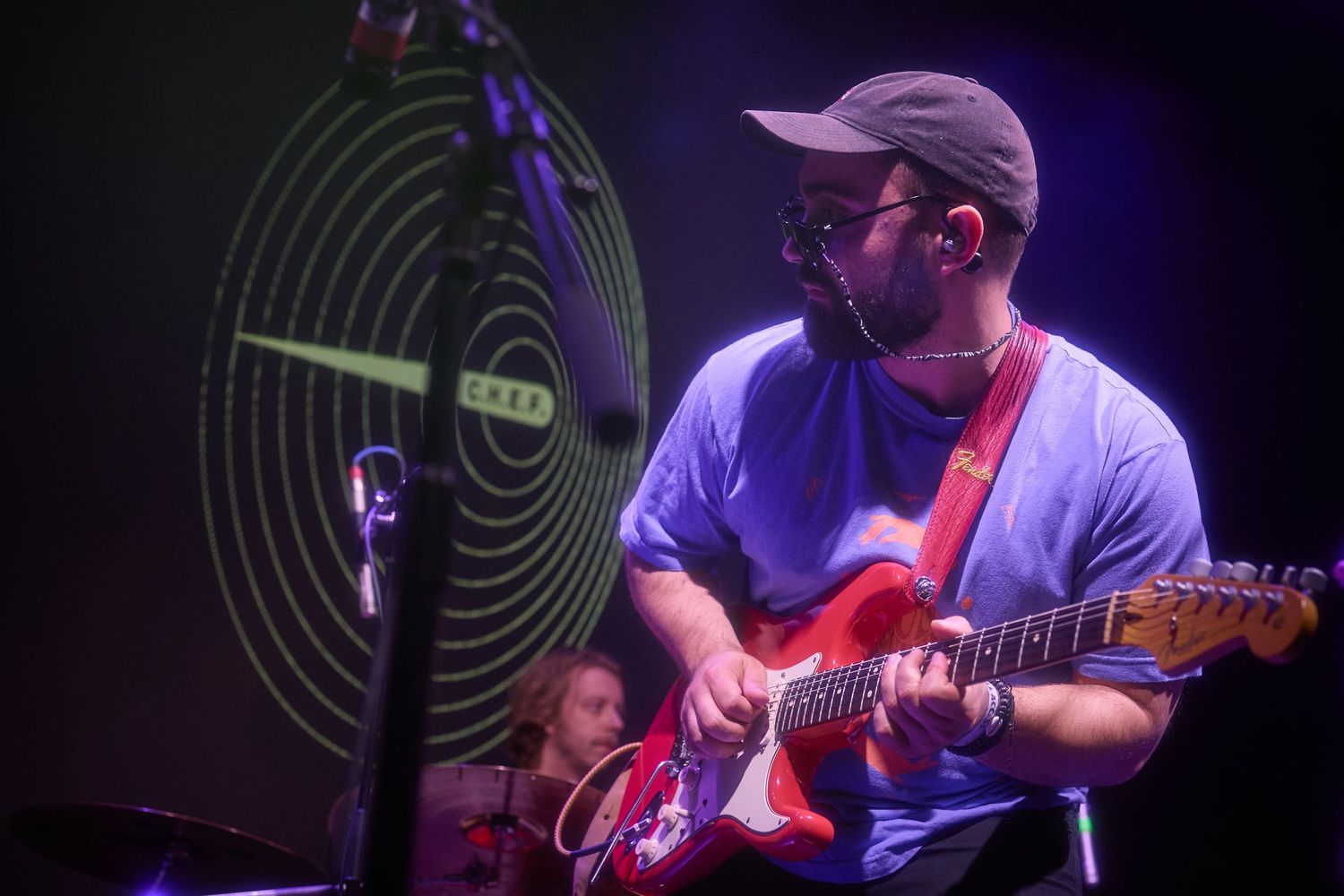 Musician performs on stage with electric guitar under purple and blue concert lighting.