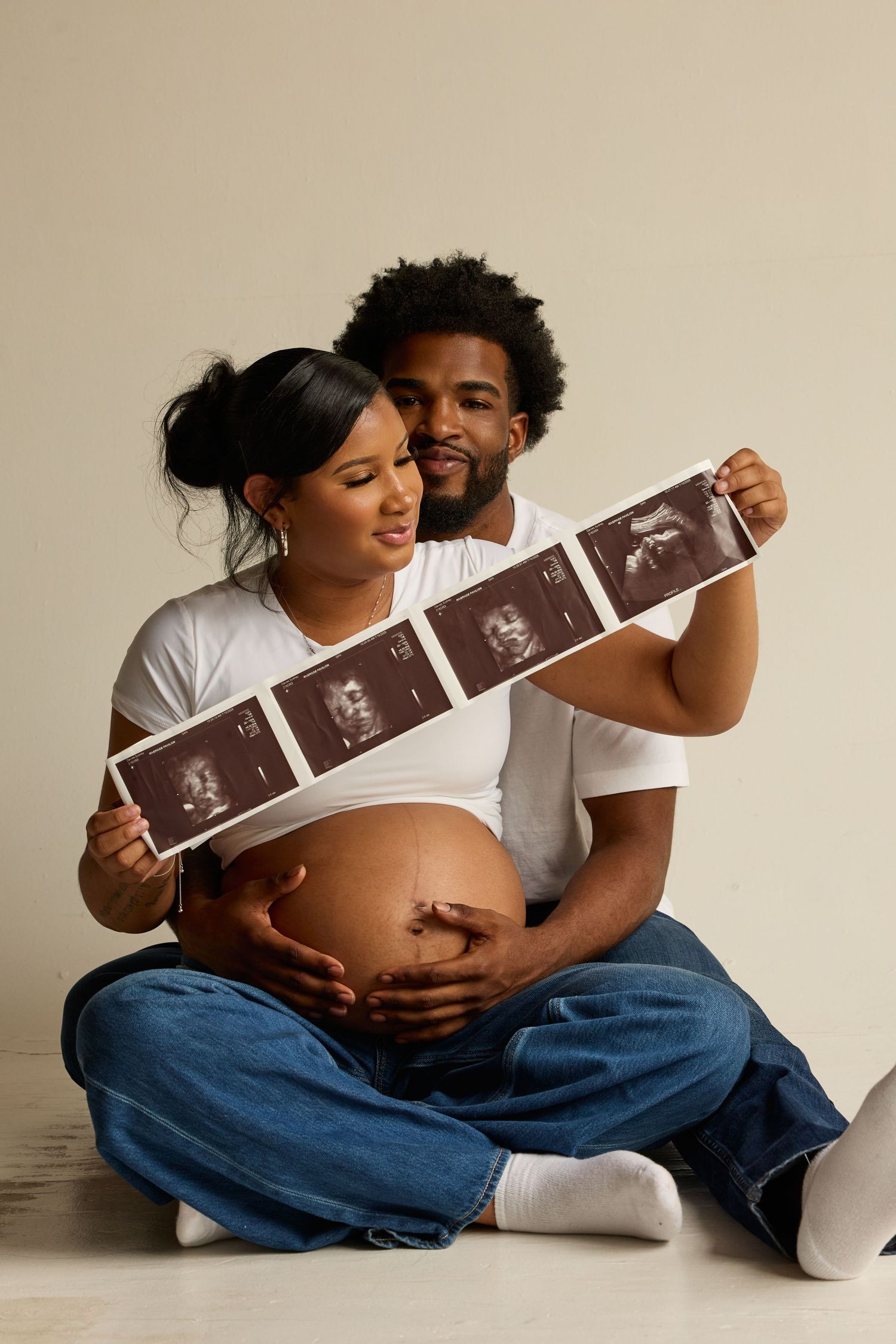 A couple sits together on the floor sharing ultrasound photos and embracing a pregnant belly with joy.