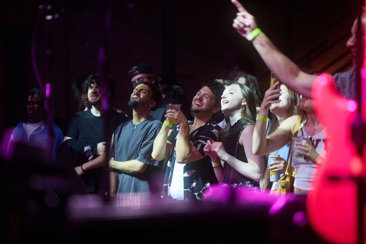 Concert crowd enjoying a nightclub performance with purple and pink lighting effects in a dark venue.