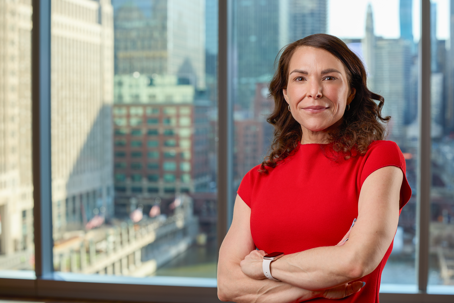 Professional in a red sleeveless top stands confidently against a city skyline backdrop in an office setting.
