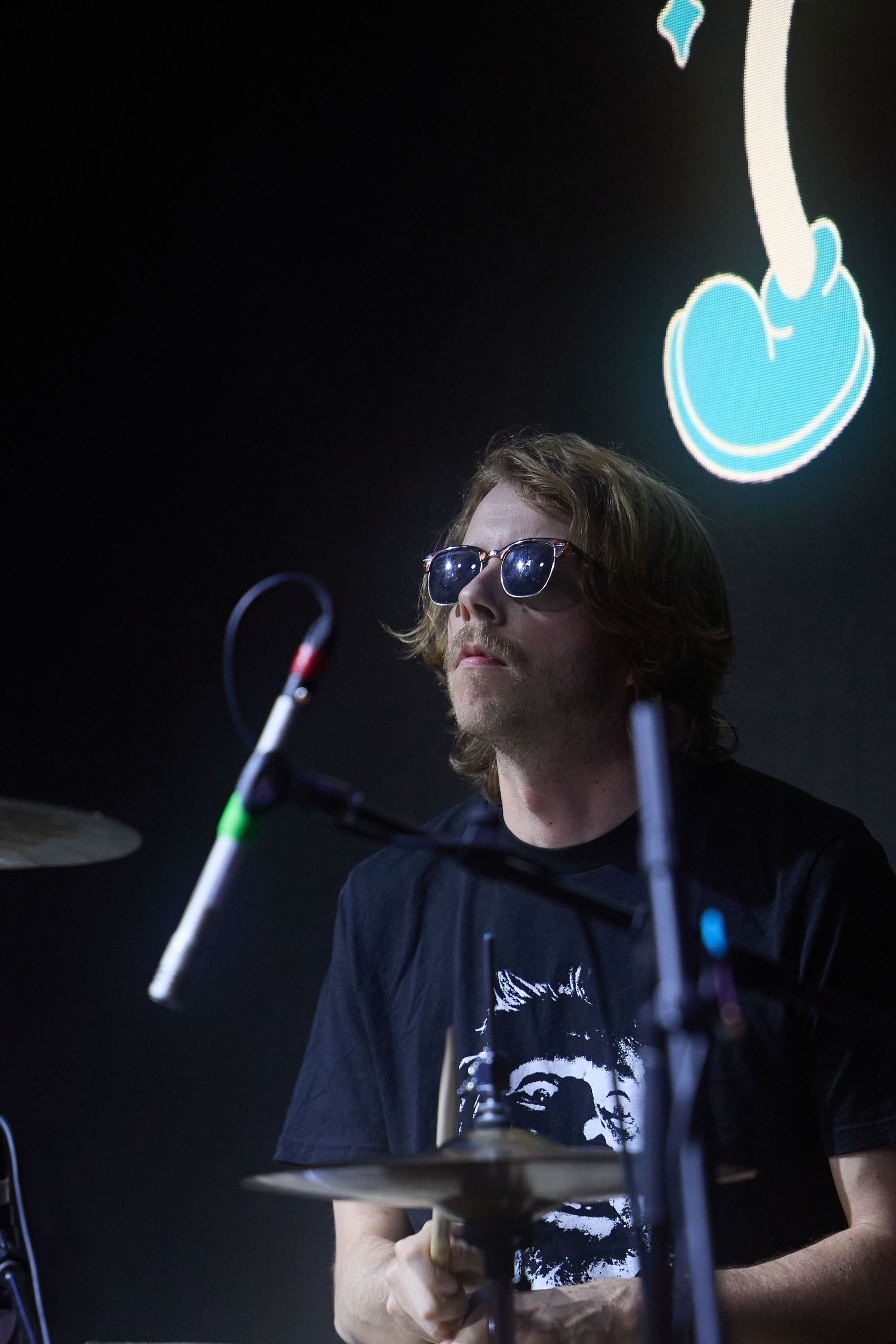 A drummer performs in low lighting with blue neon accents visible in the background.