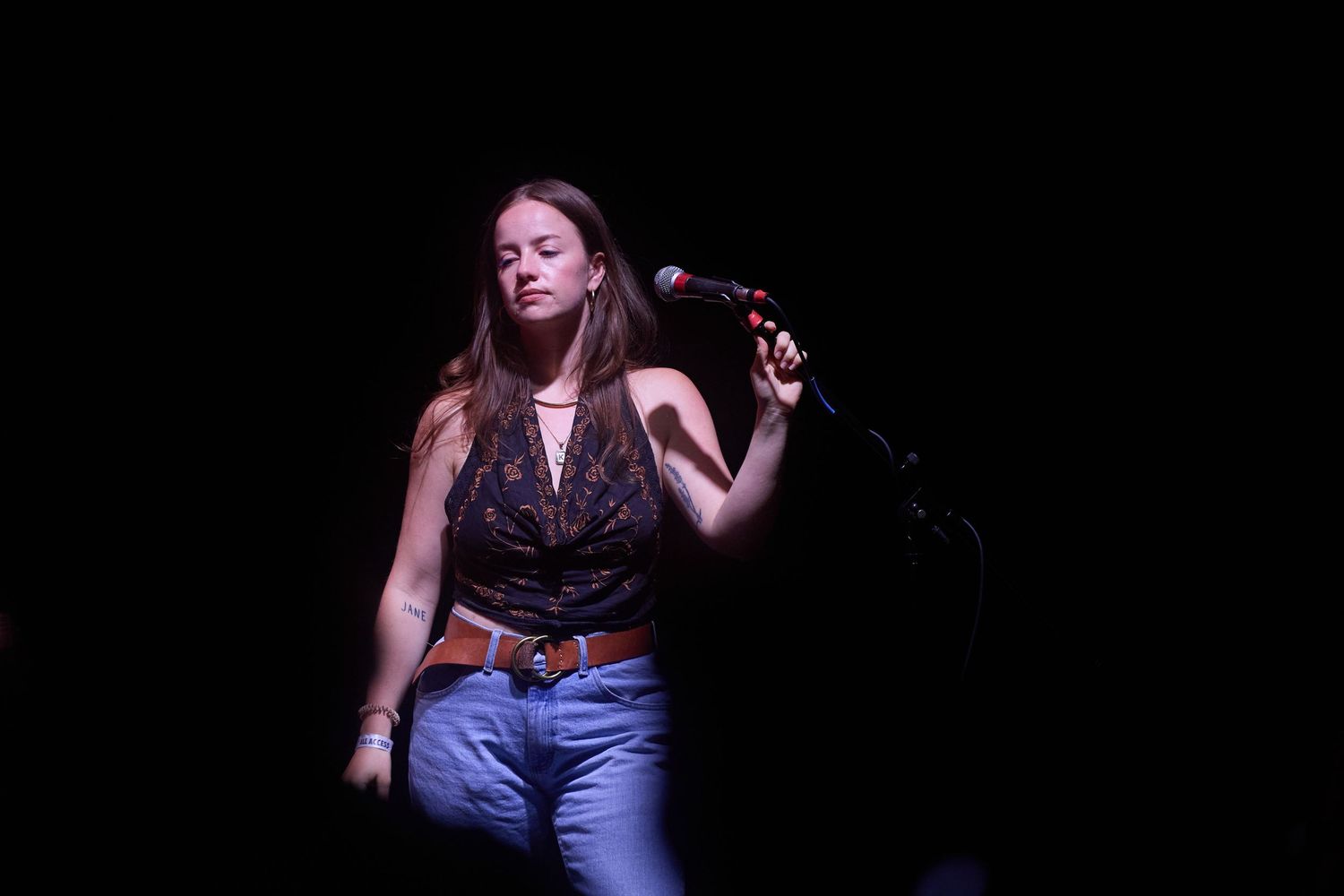 A performer in a blue dress gestures expressively on a dark stage during a theatrical performance.