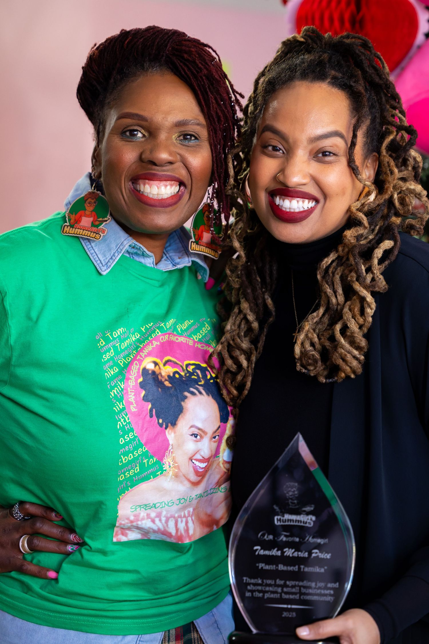 Two women smile brightly while posing together, one wearing a green shirt and one in black, holding a glass award trophy.