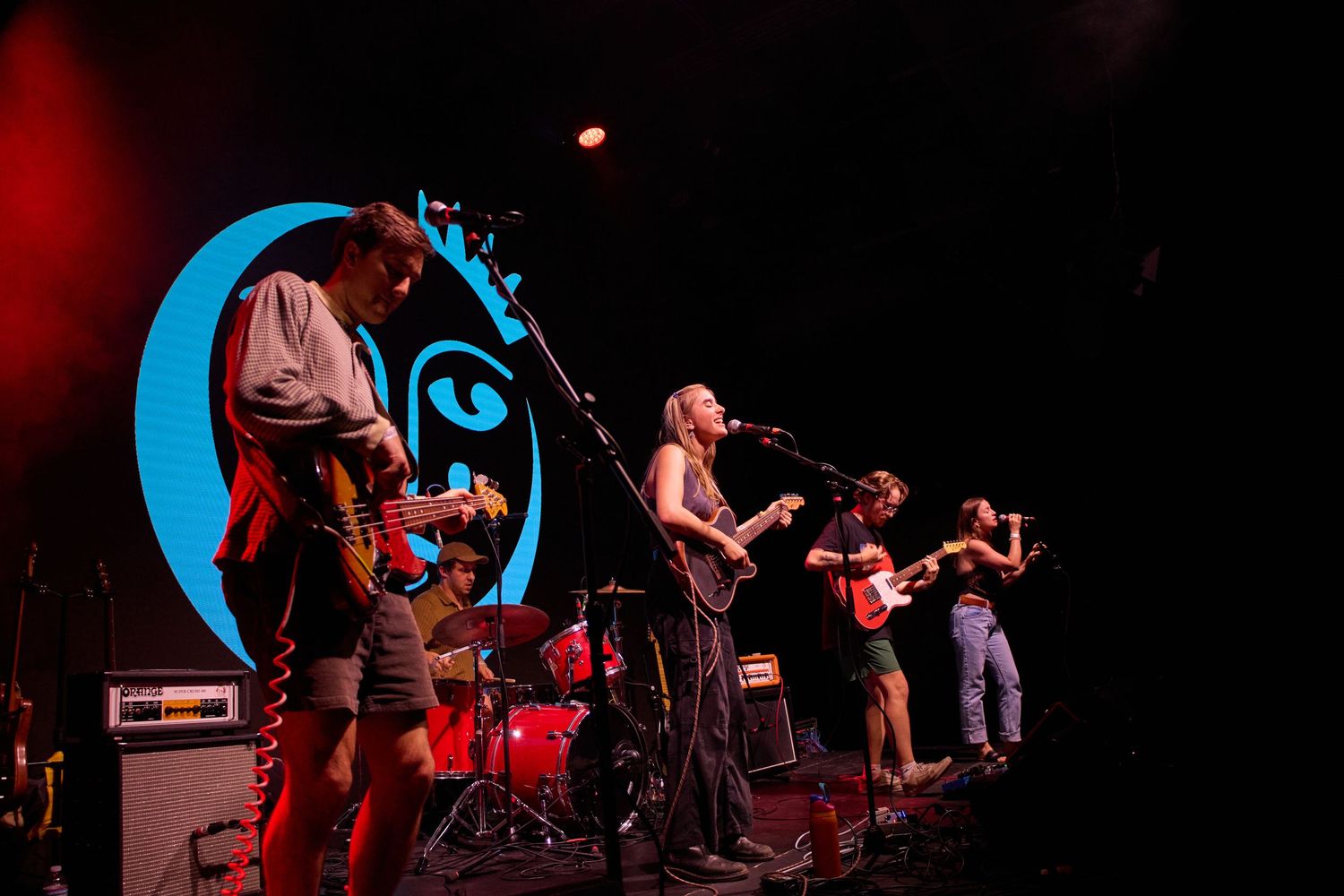 Musicians perform on stage at a concert with dramatic red and blue lighting in the background.