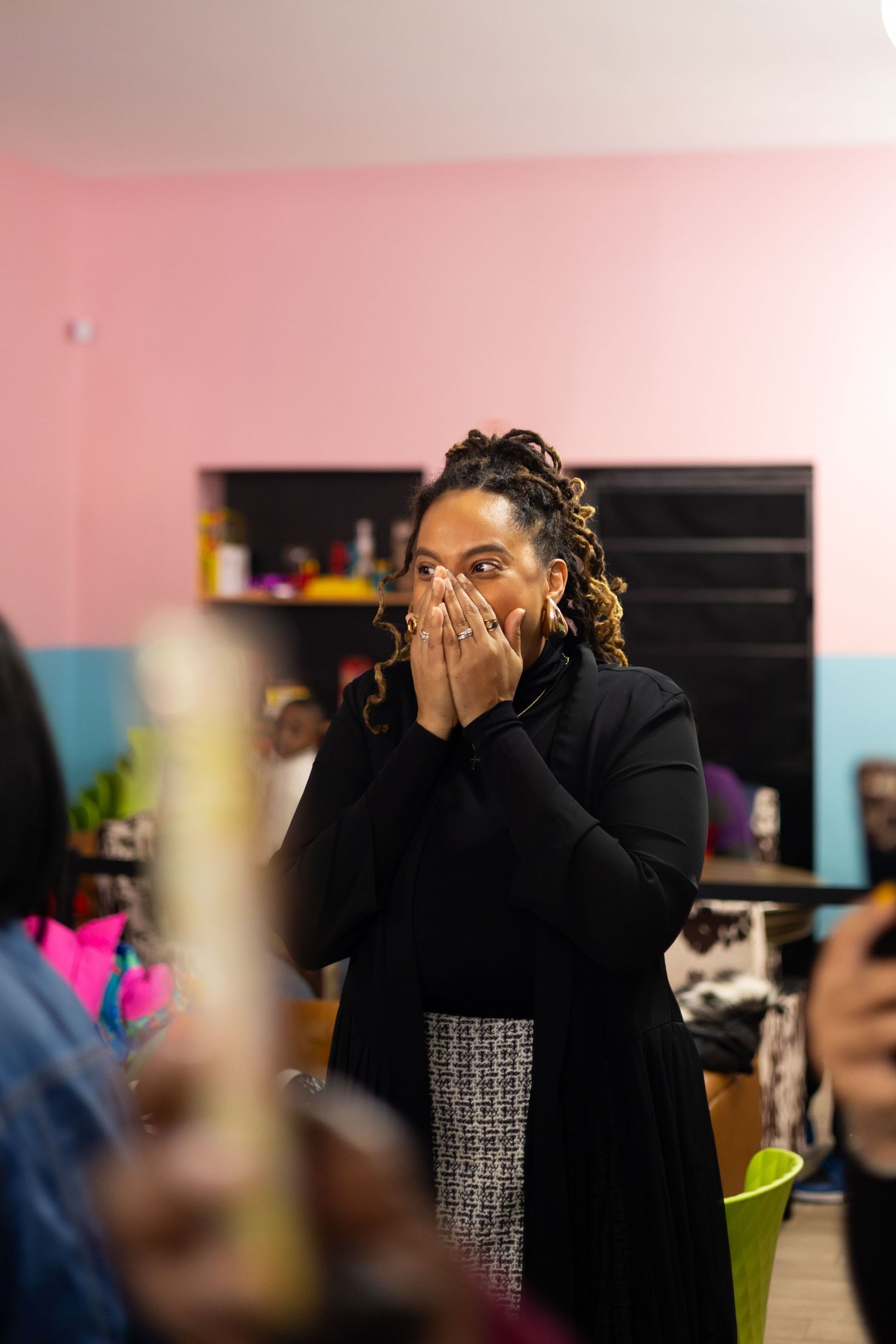 A person wearing a black coat and face mask stands in a room with blurred foreground elements and pink walls.