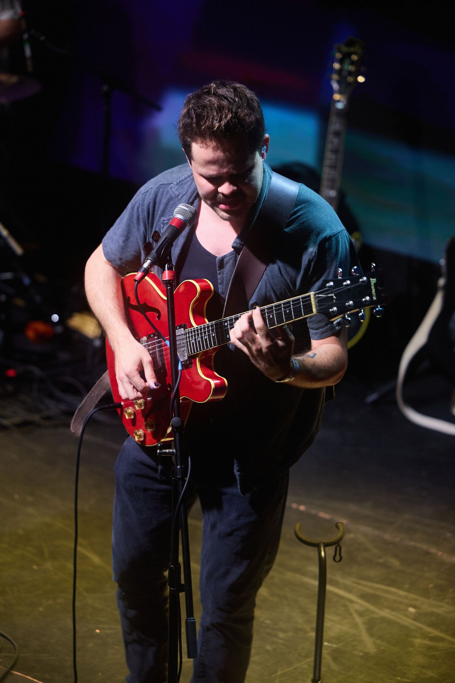 A musician performs on stage with a red electric guitar in a dimly lit venue.