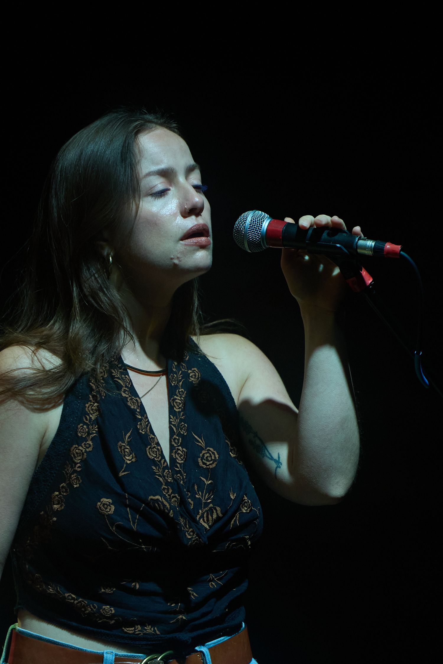 A singer performs in a dark venue wearing a black and gold patterned halter top against a dramatic black background.