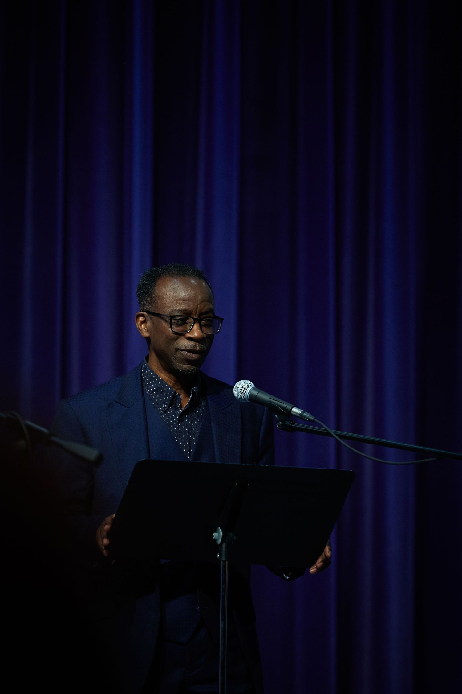 A speaker stands at a podium with a microphone against a dark blue curtain backdrop during a performance.