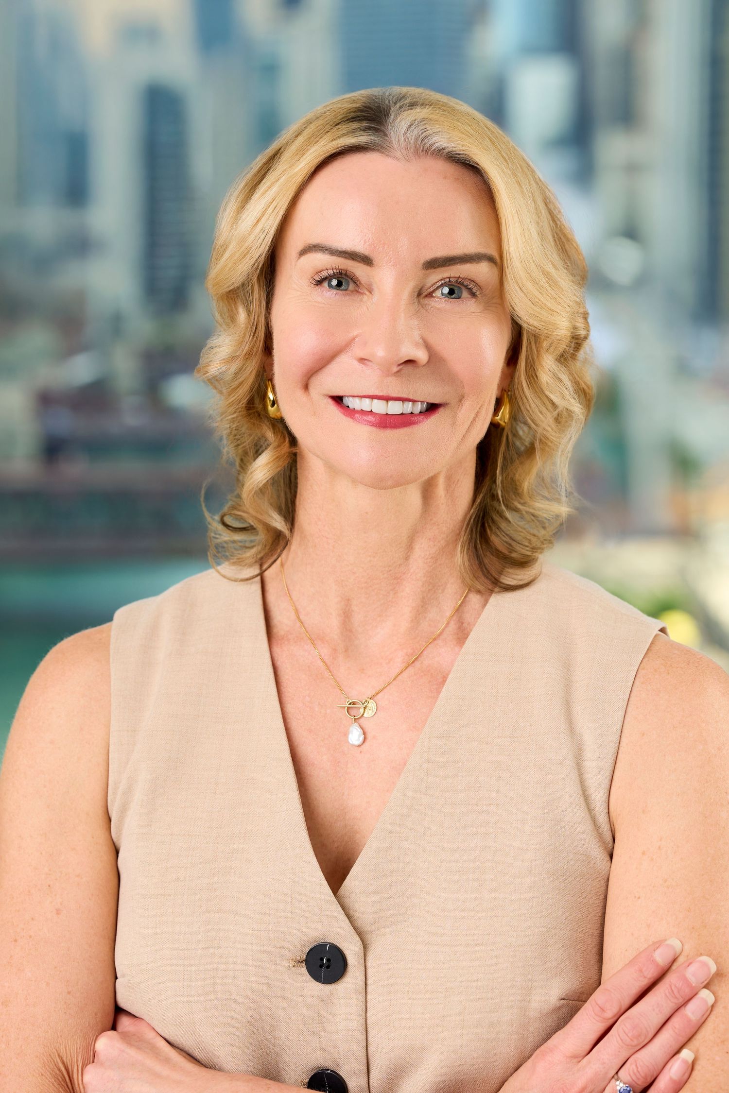 Professional headshot of a woman in a beige sleeveless top smiling confidently against a blurred city background.