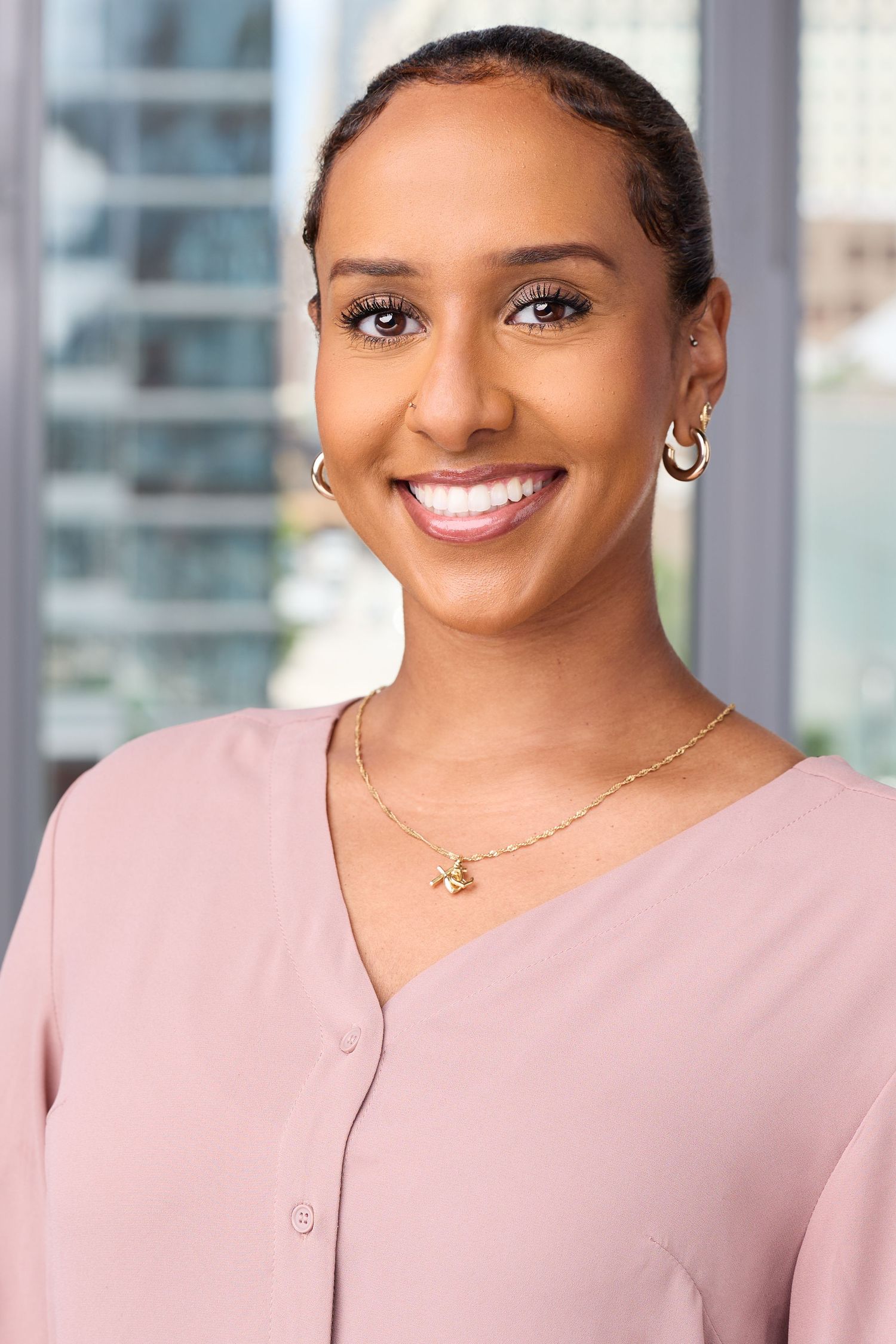 Professional headshot of someone wearing a soft pink blouse and smiling warmly at the camera in an office setting.