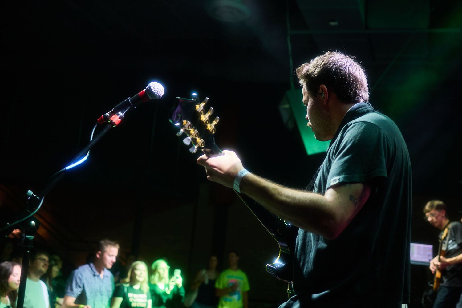 Musicians perform on a dark stage with green lighting and silhouetted figures visible in the background.