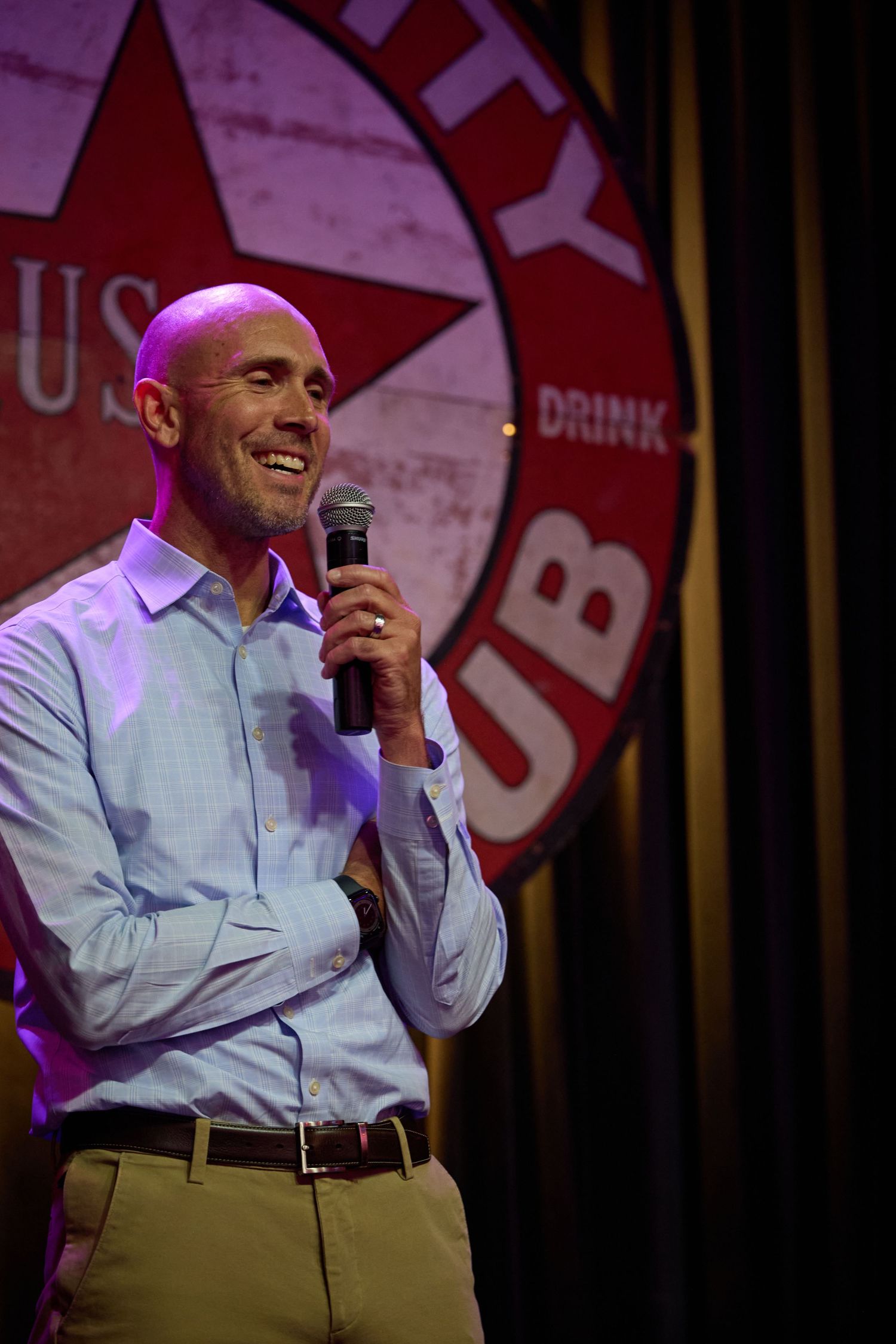 A speaker in a light blue dress shirt and khaki pants smiles while holding a microphone on stage.
