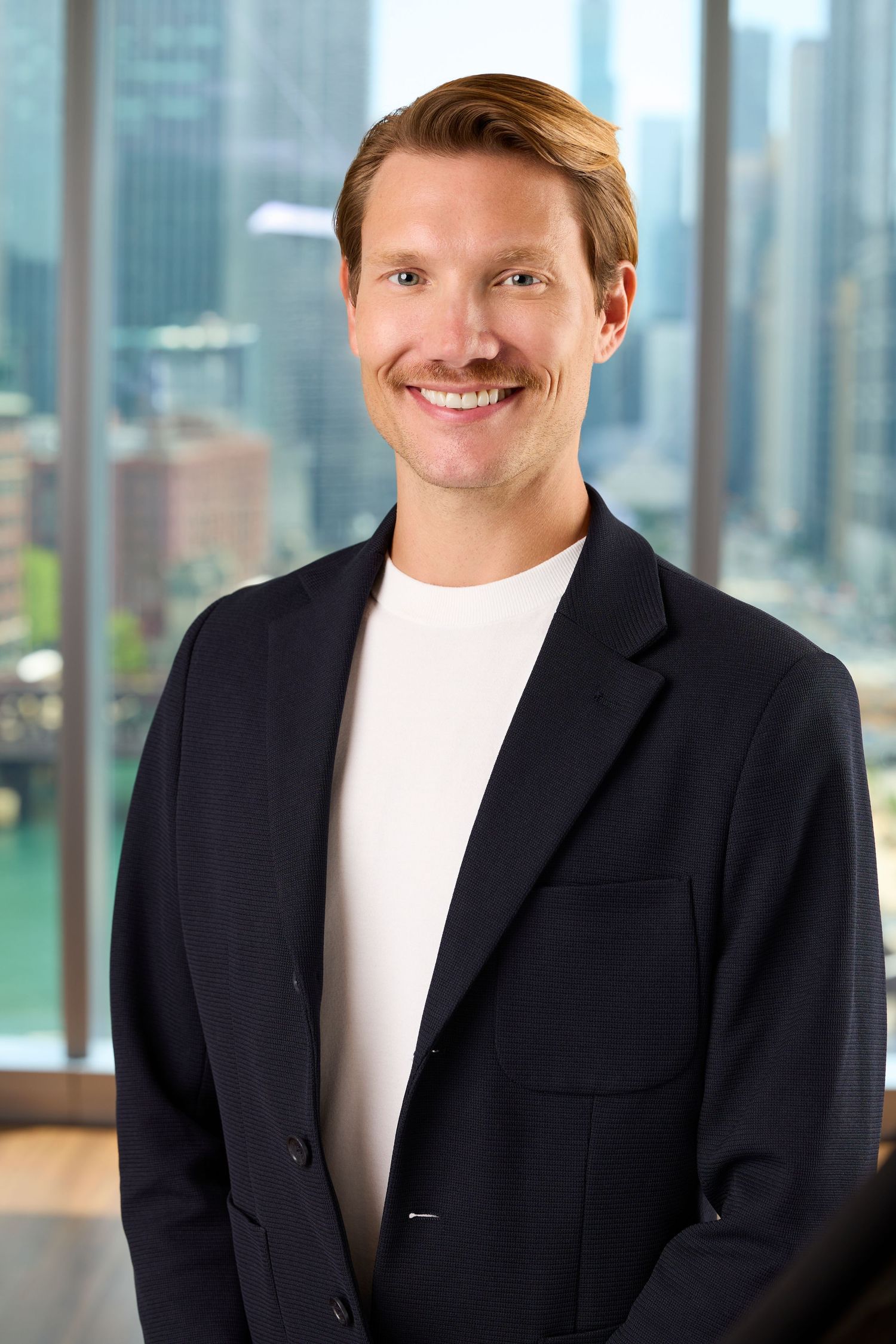 Professional headshot of someone in a dark suit jacket and white shirt smiling against a blurred office background.