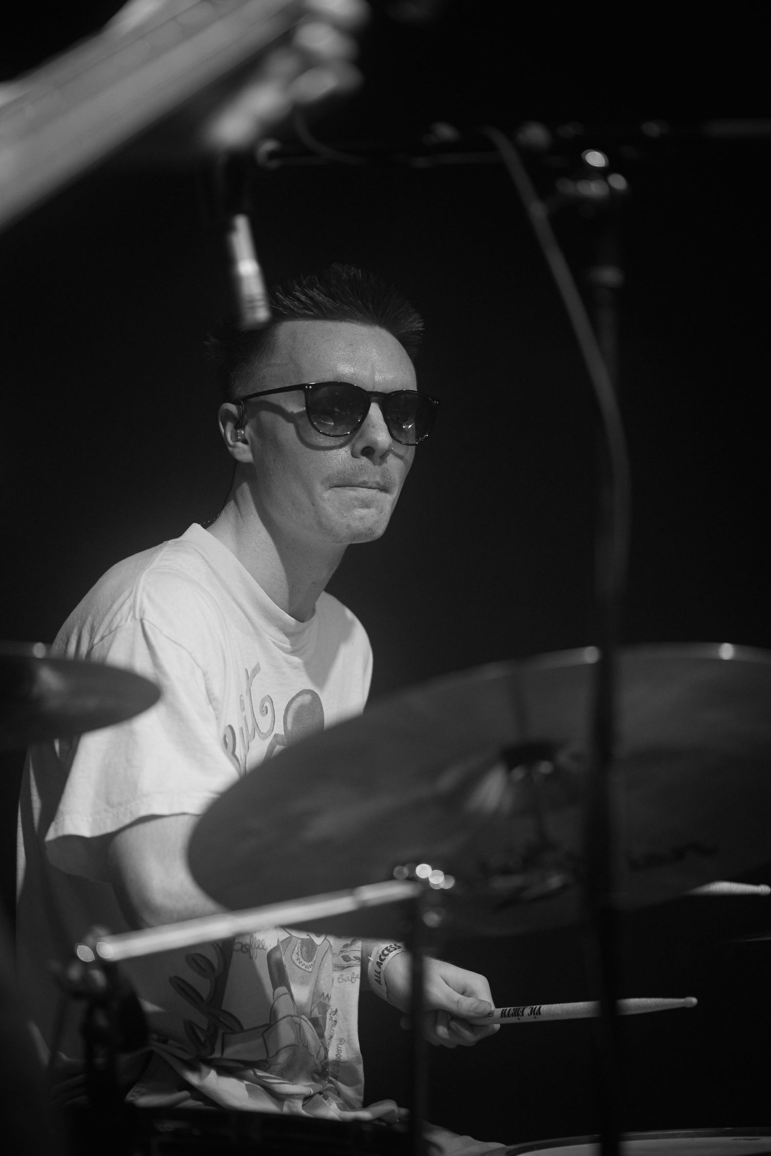 Drummer wearing sunglasses and a white t-shirt performs on stage in black and white lighting.