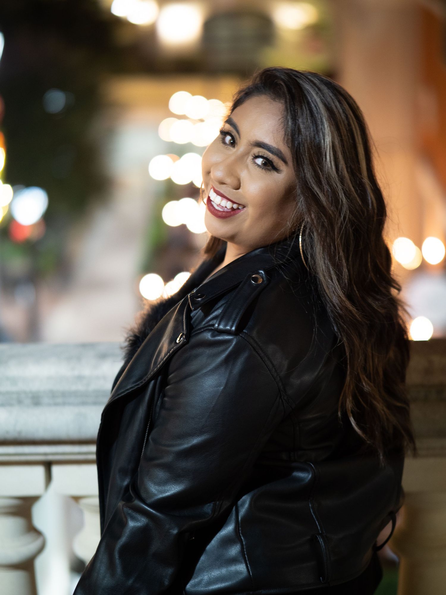 Stylish woman in black leather jacket poses against blurred city lights at night for fashion photography series.