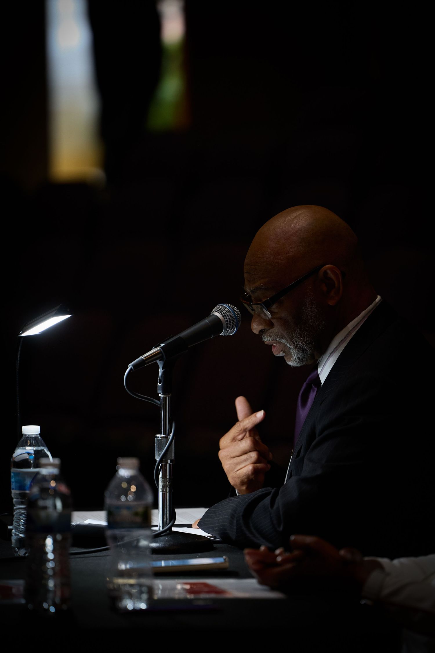 A person in a dark suit speaks into a microphone at a conference table with water bottles visible in low lighting.