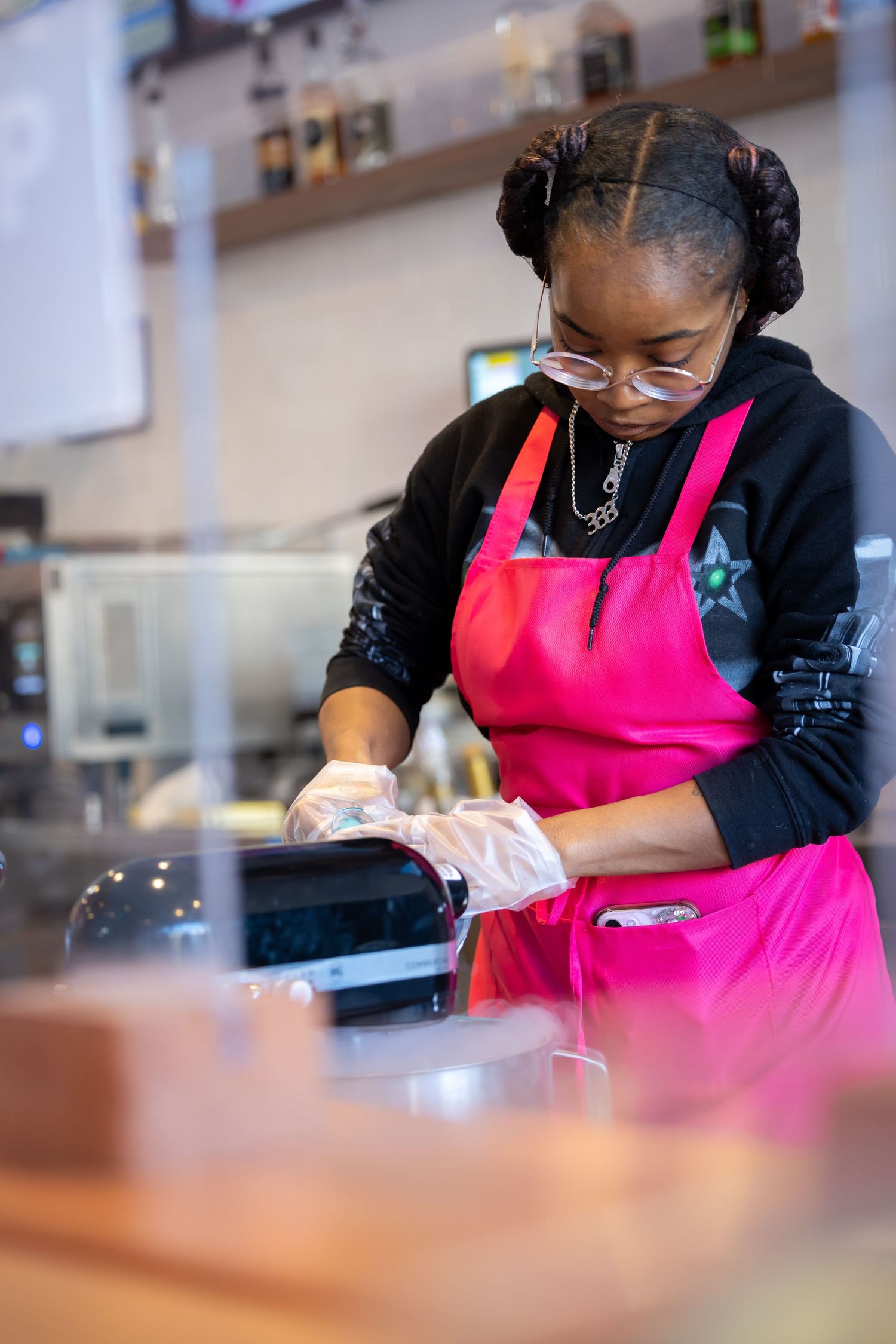 A person in a bright pink apron and black shirt carefully works on a detailed task at a wooden counter.