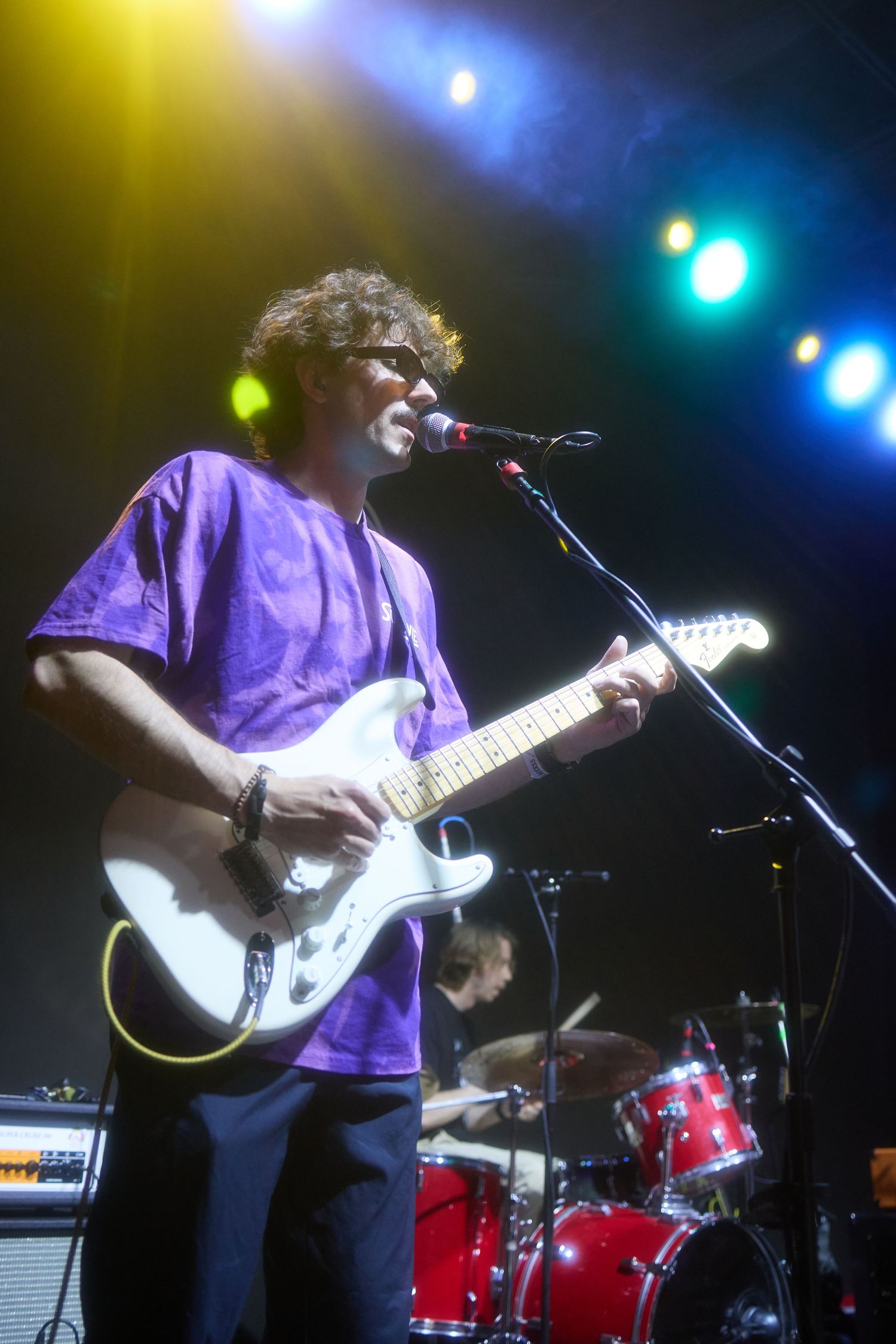A guitarist performs on stage with a white electric guitar under blue and green stage lighting.