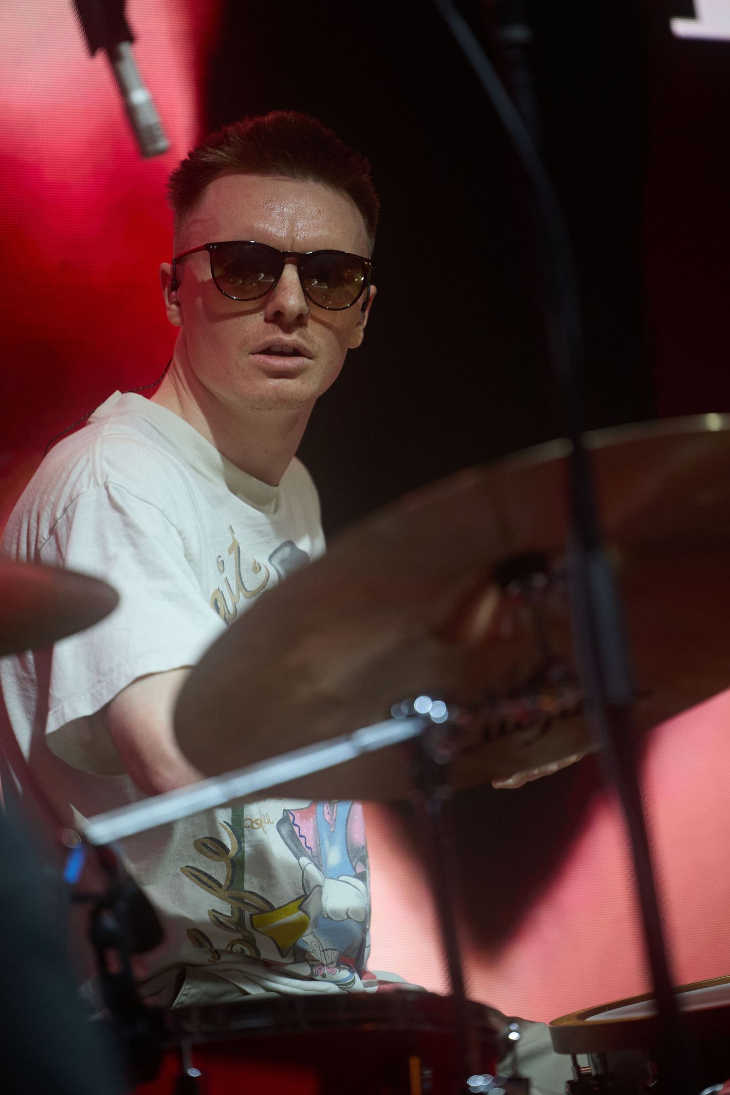 A drummer performs on stage in a white t-shirt under dramatic red lighting during a live music performance.