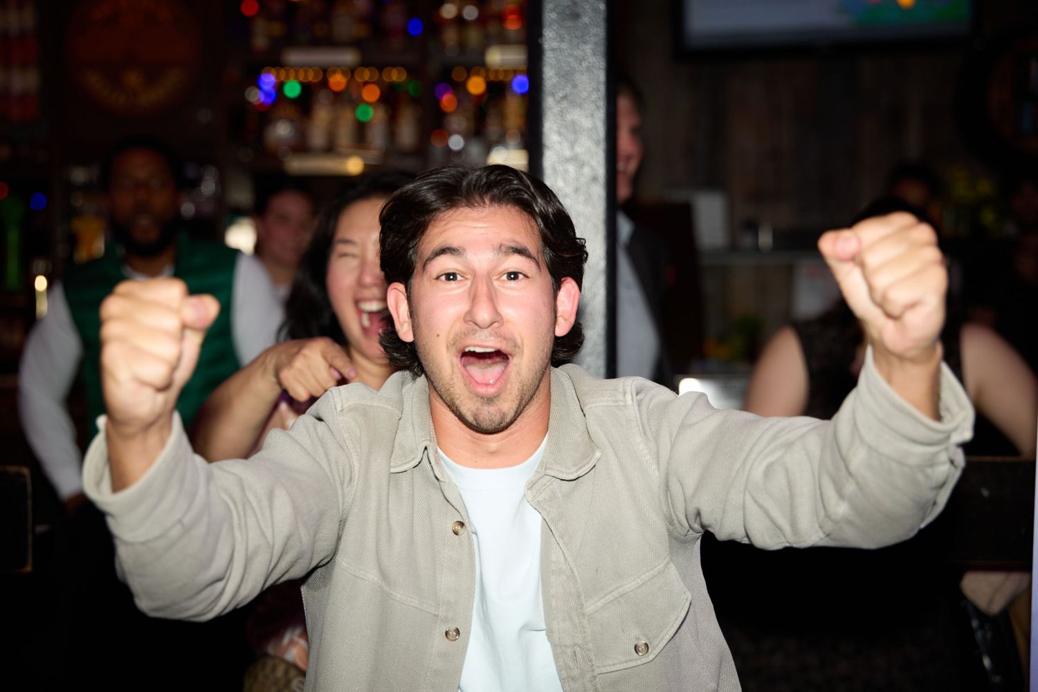 Person gestures excitedly with raised arms while wearing a light colored shirt in a dimly lit setting.