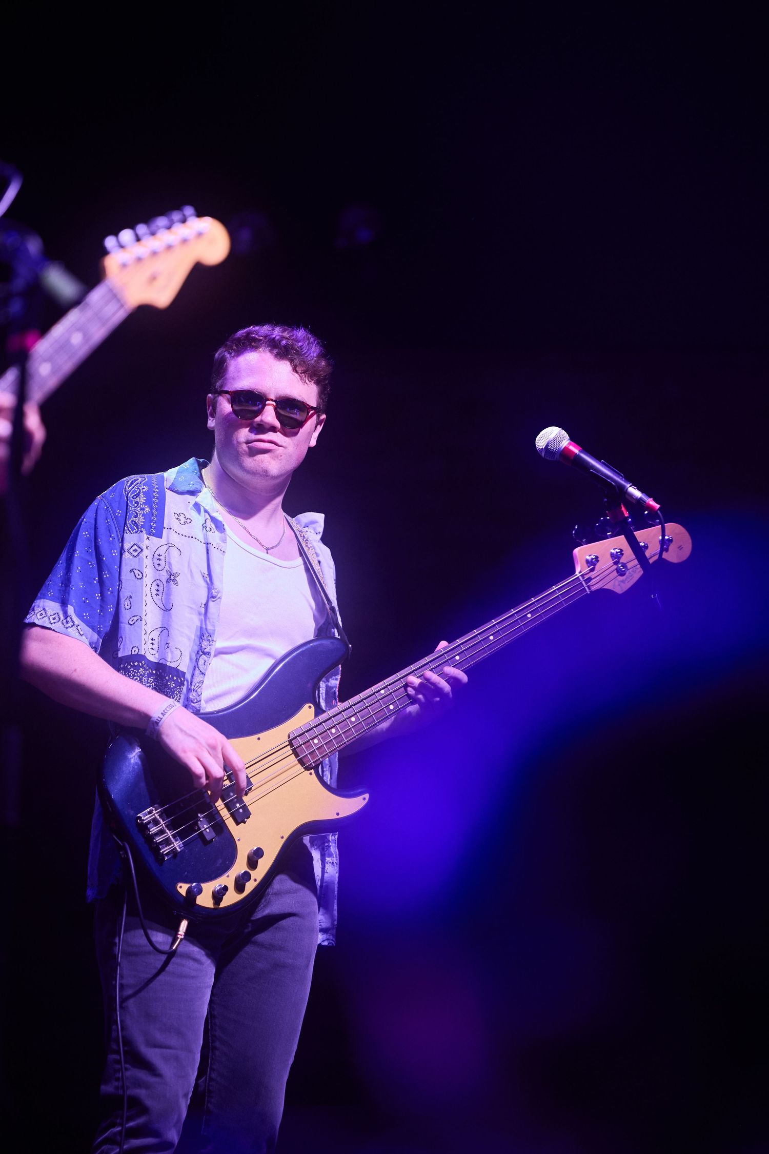 Bass guitarist wearing sunglasses performs on stage with a cream colored bass guitar under purple stage lighting.