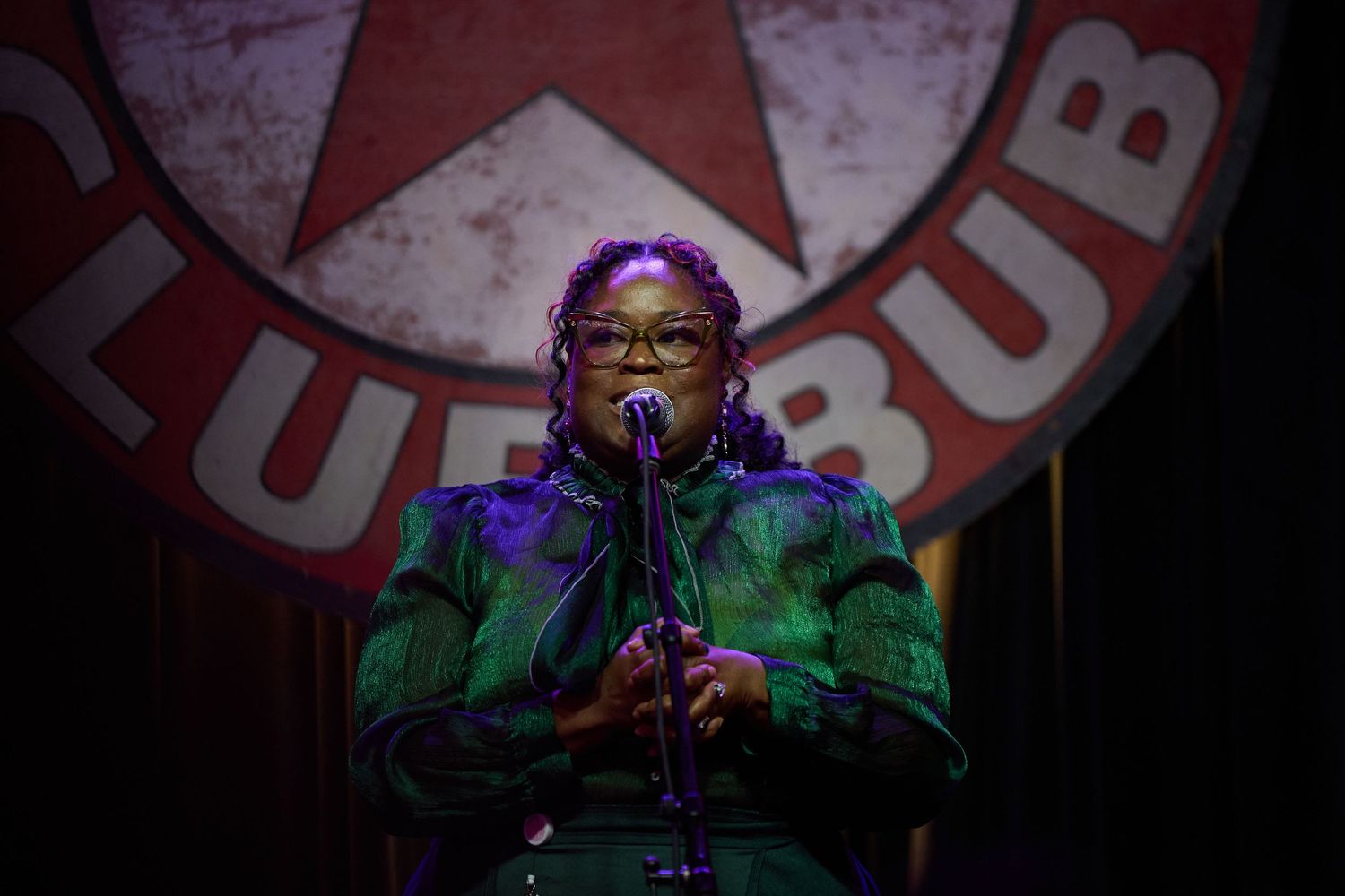 A performer in a green jacket sings into a microphone on stage with red lighting and signage in the background.