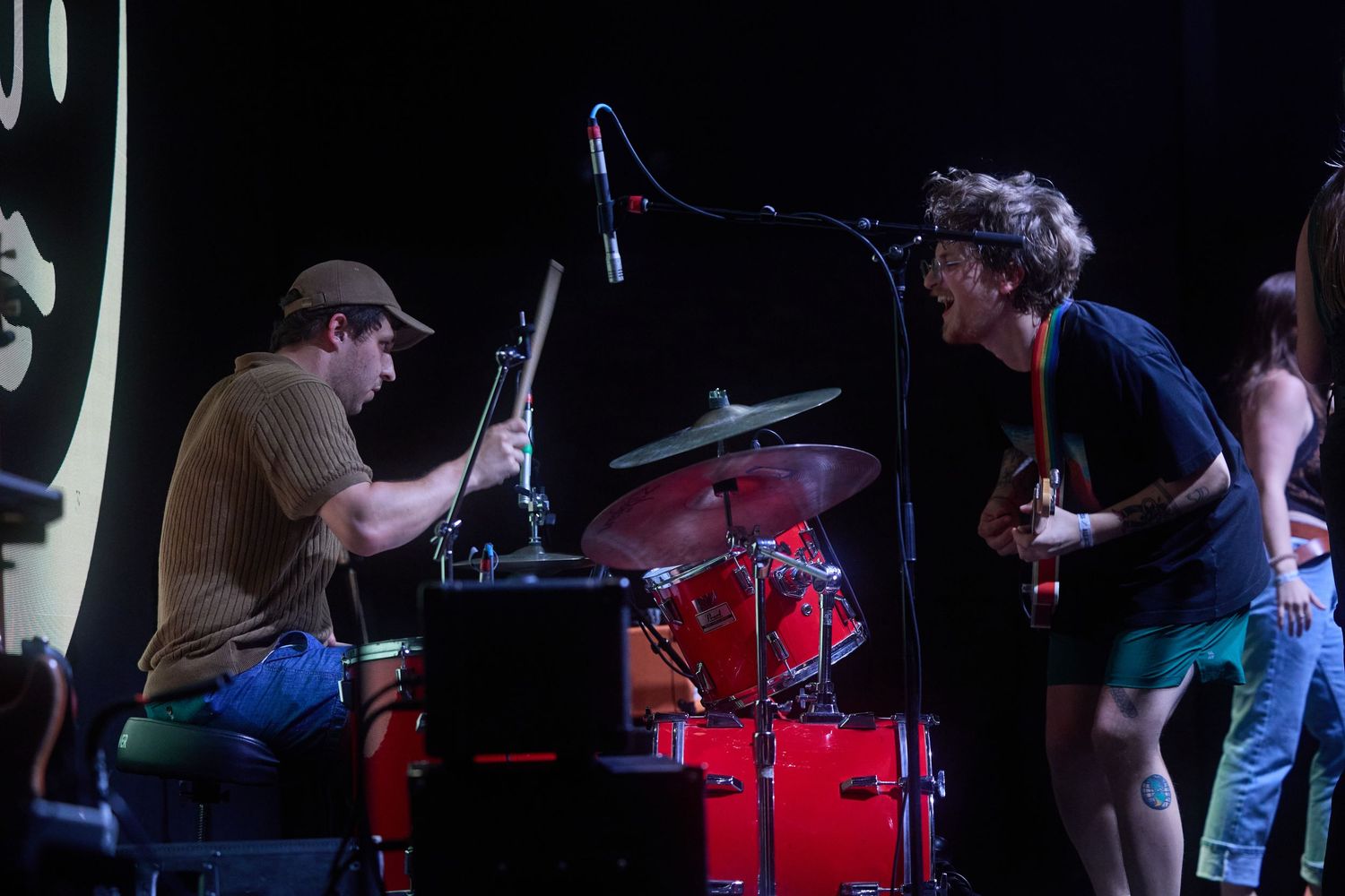 Musicians perform on stage with drums and instruments during a dark concert setting.