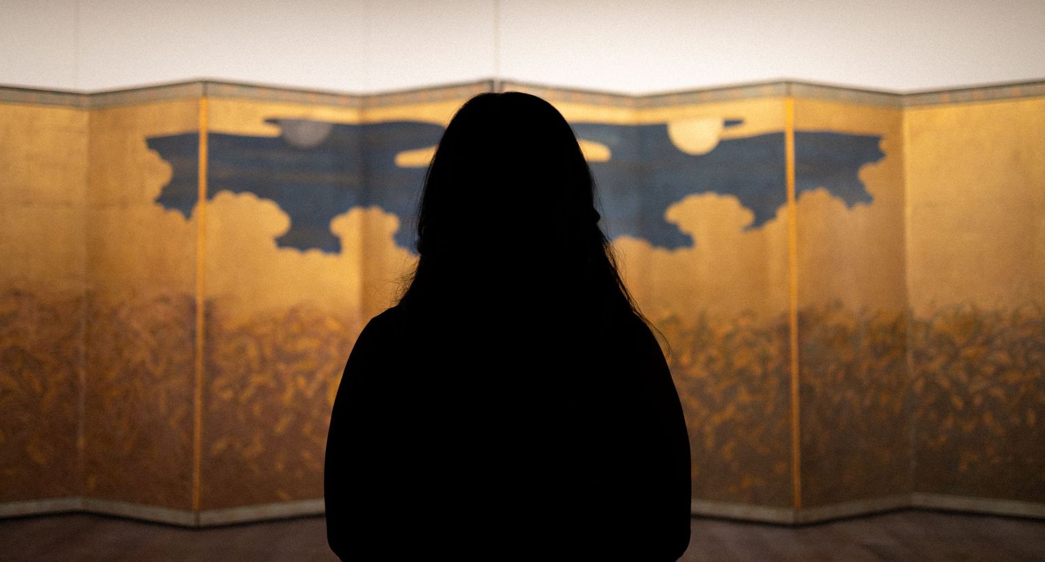 A silhouetted figure sits in front of a golden Japanese folding screen display in a dimly lit room.