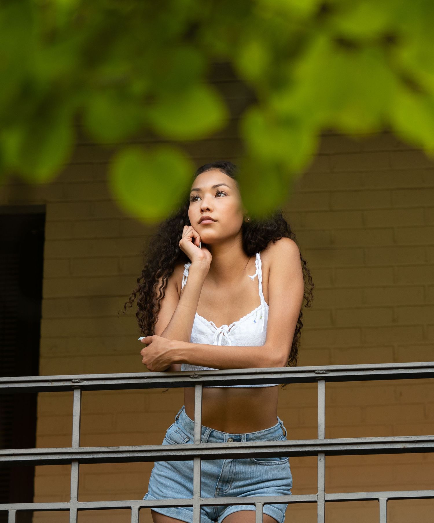 Person in denim shorts leans thoughtfully on a metal railing against a green leafy background.