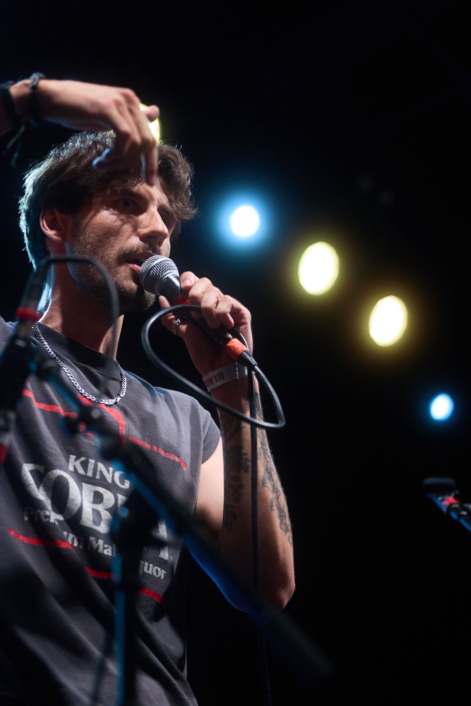 Concert performer in a dark room sings into microphone on stage with blue and yellow stage lights in background.