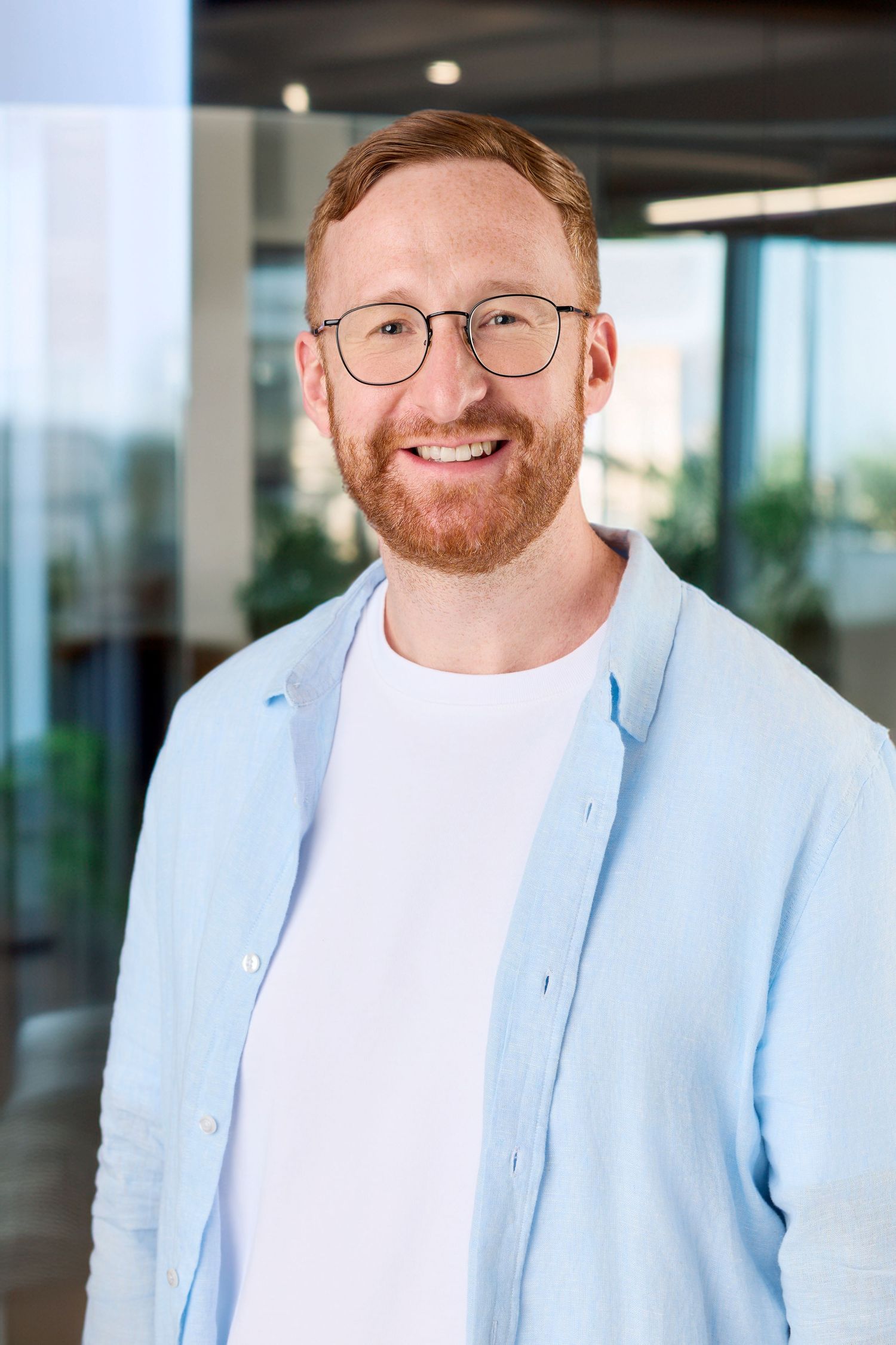 Professional headshot of someone wearing glasses and a light blue blazer with white shirt in an office setting.