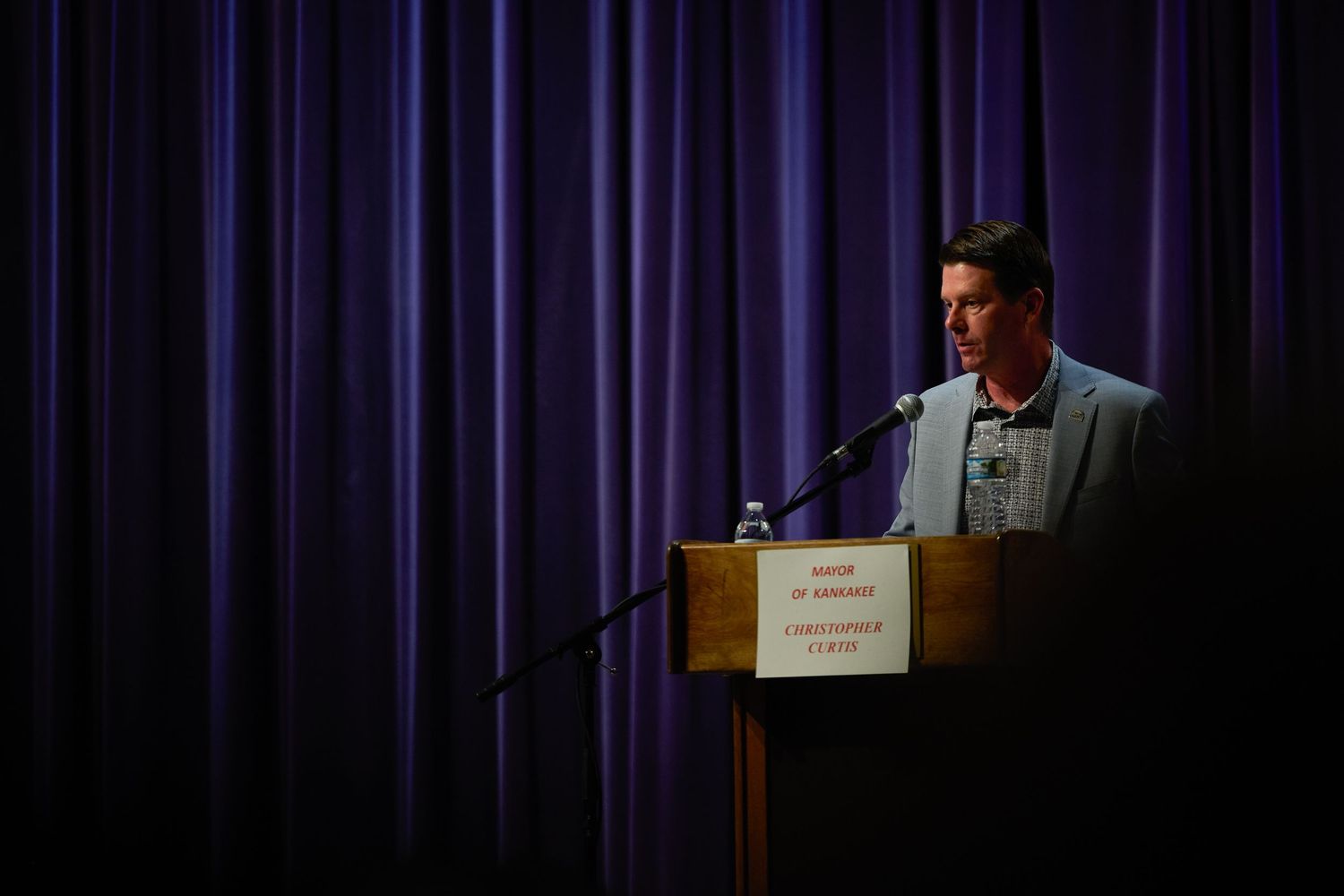 A speaker stands at a podium on stage with purple curtains in the background during a presentation.