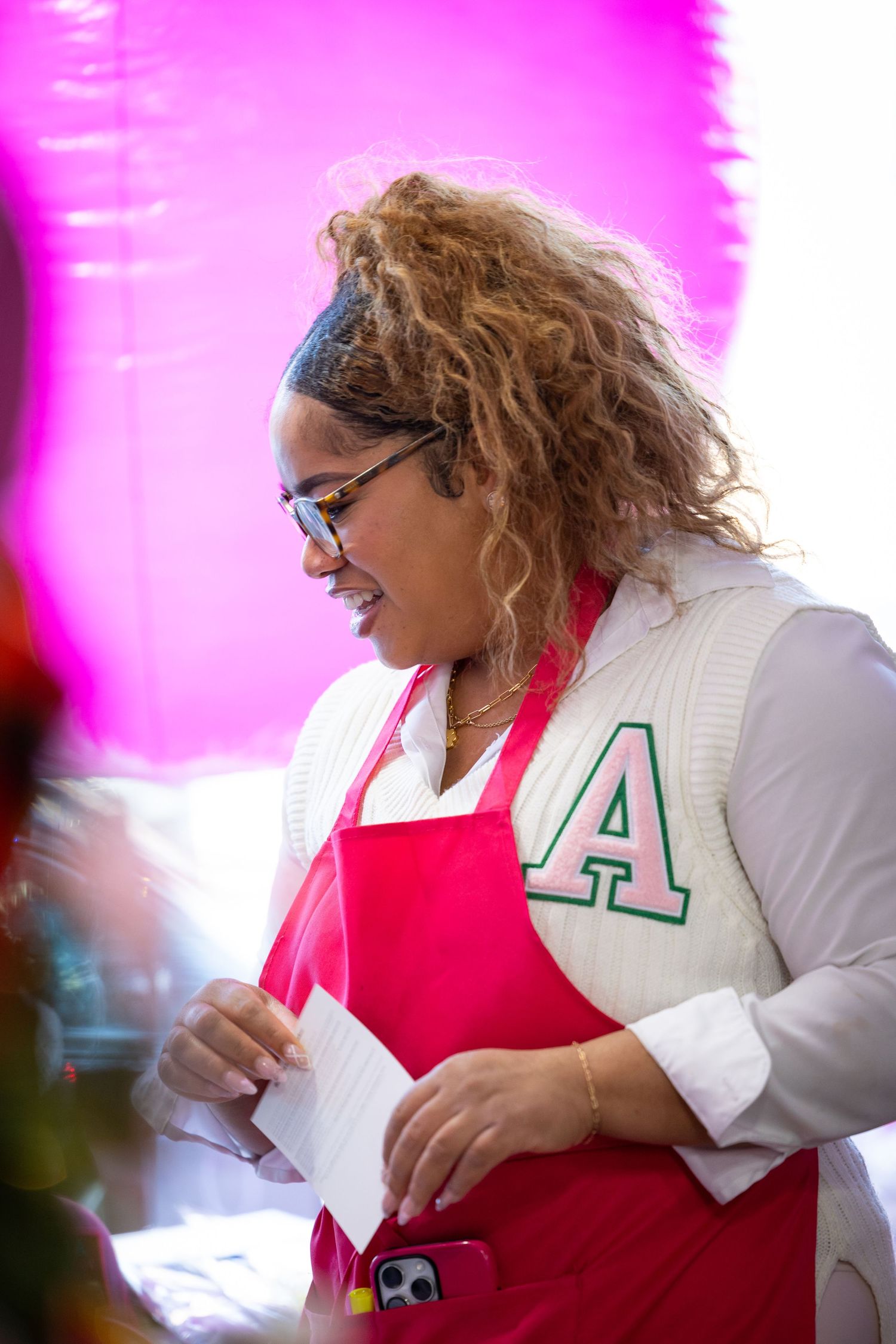 A person in a white cardigan and pink apron writes while sitting at a desk with a pink-lit background.