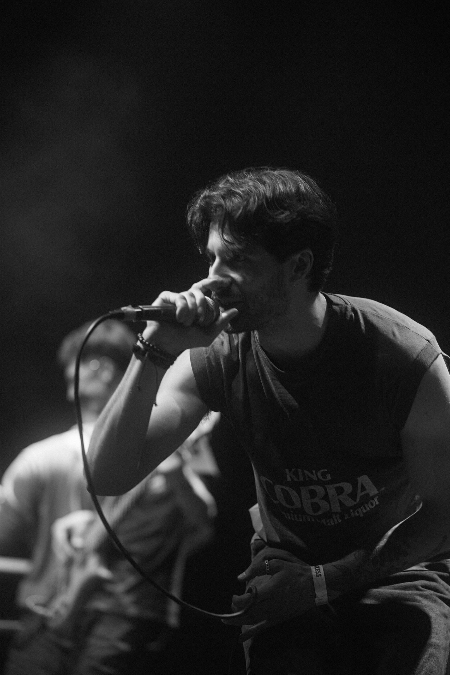 A performer in a casual t-shirt sings passionately into a microphone on stage in black and white.