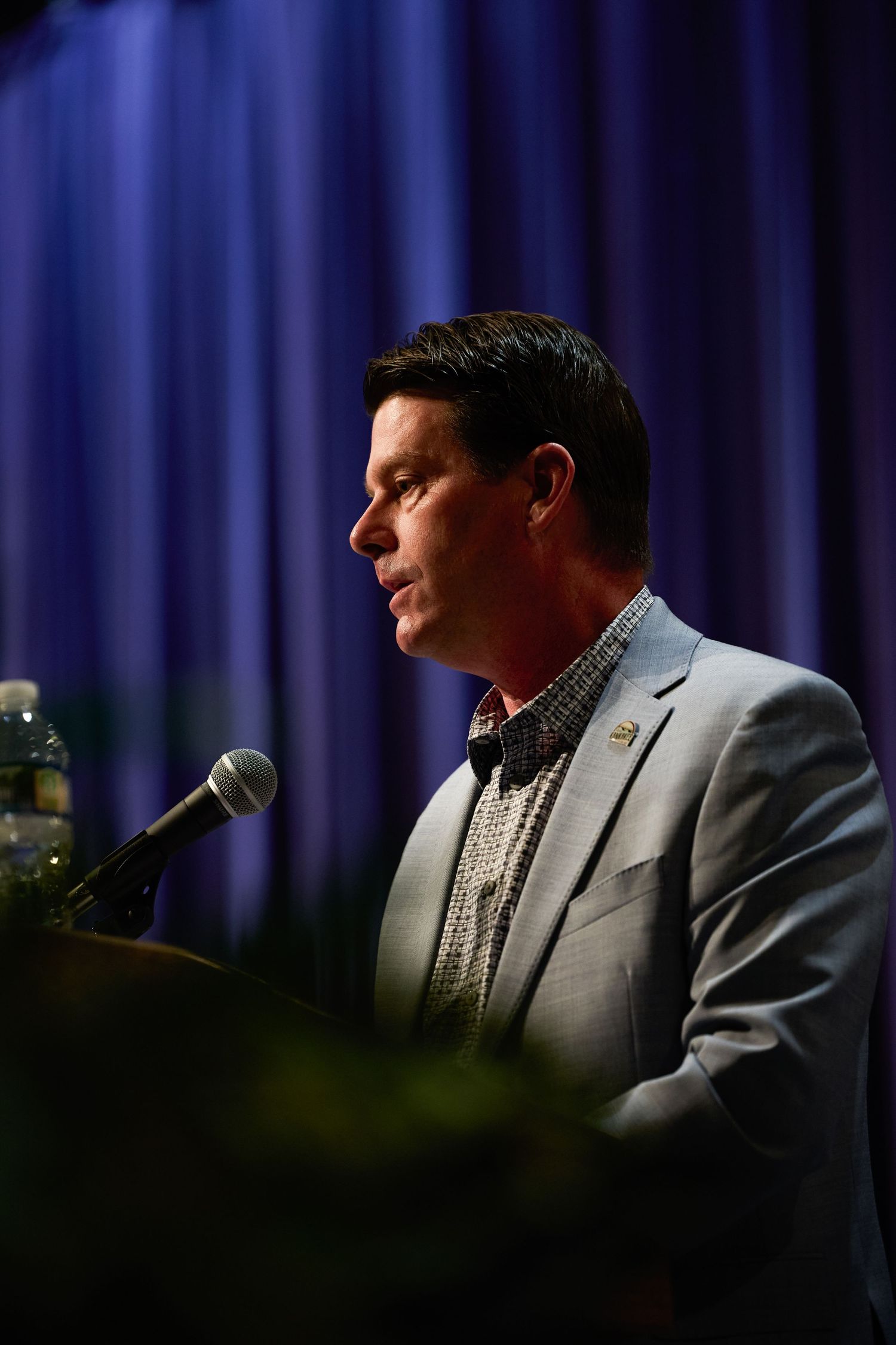 A person in a gray suit speaks at a podium with a microphone against a dark blue curtained backdrop.