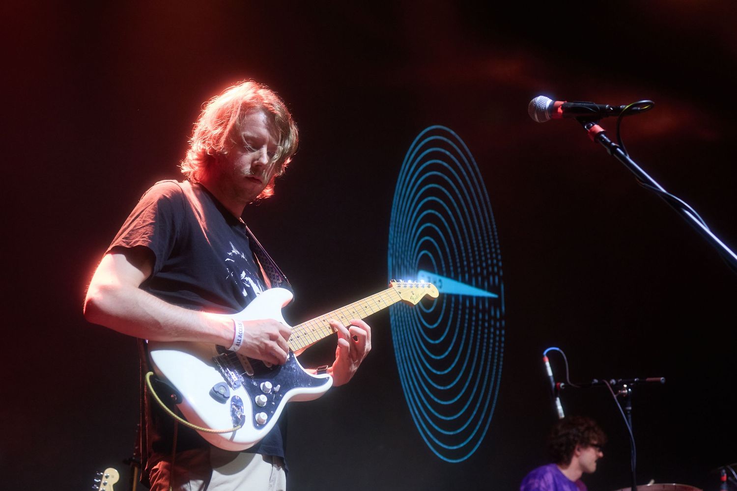 A guitarist performs on stage with a glowing red light effect and illuminated spiral backdrop during a live concert performance.