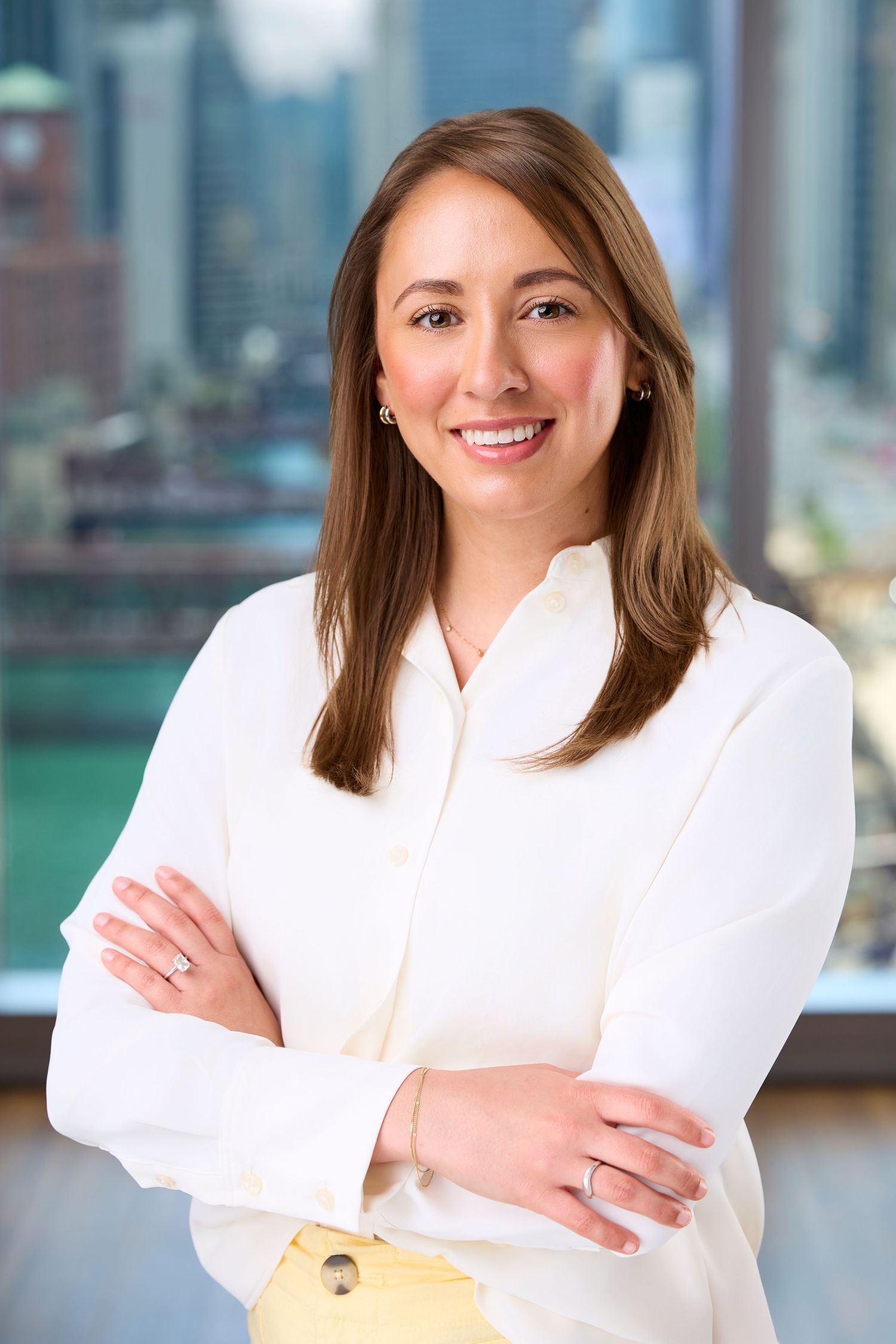 Professional headshot of a smiling businesswoman in a white blouse standing with arms crossed in front of city windows.