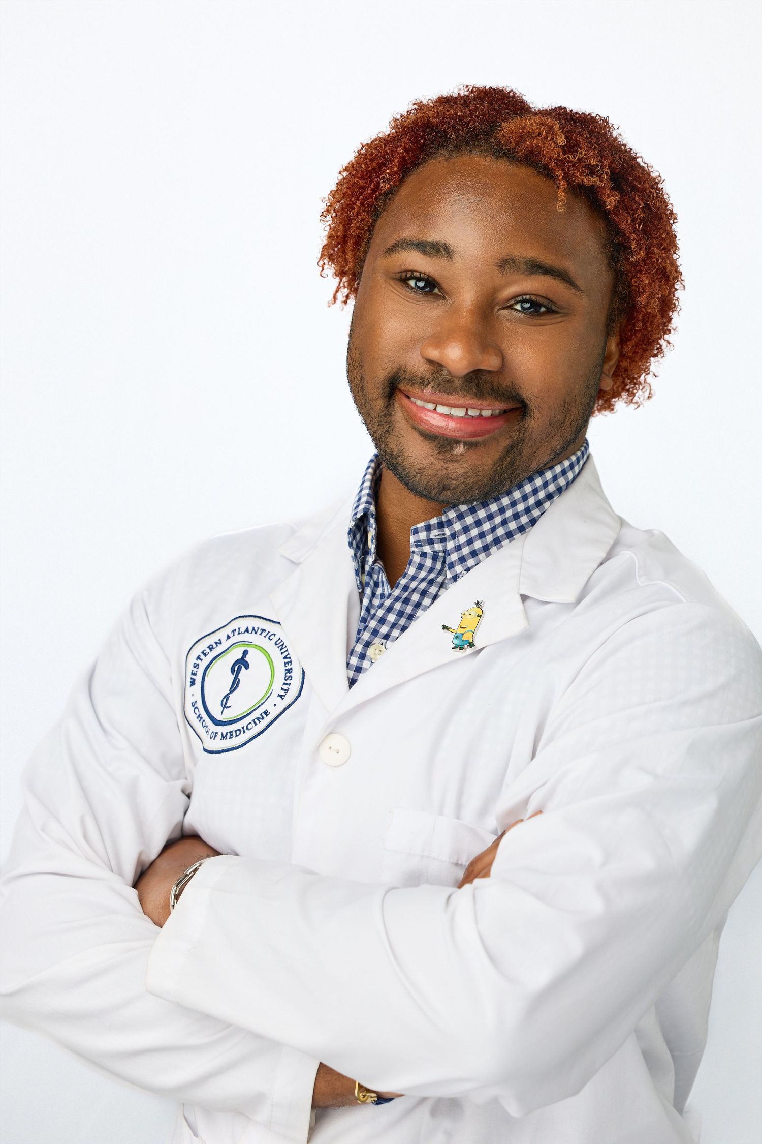 Healthcare professional in white lab coat and checkered shirt with arms crossed smiling against white background.