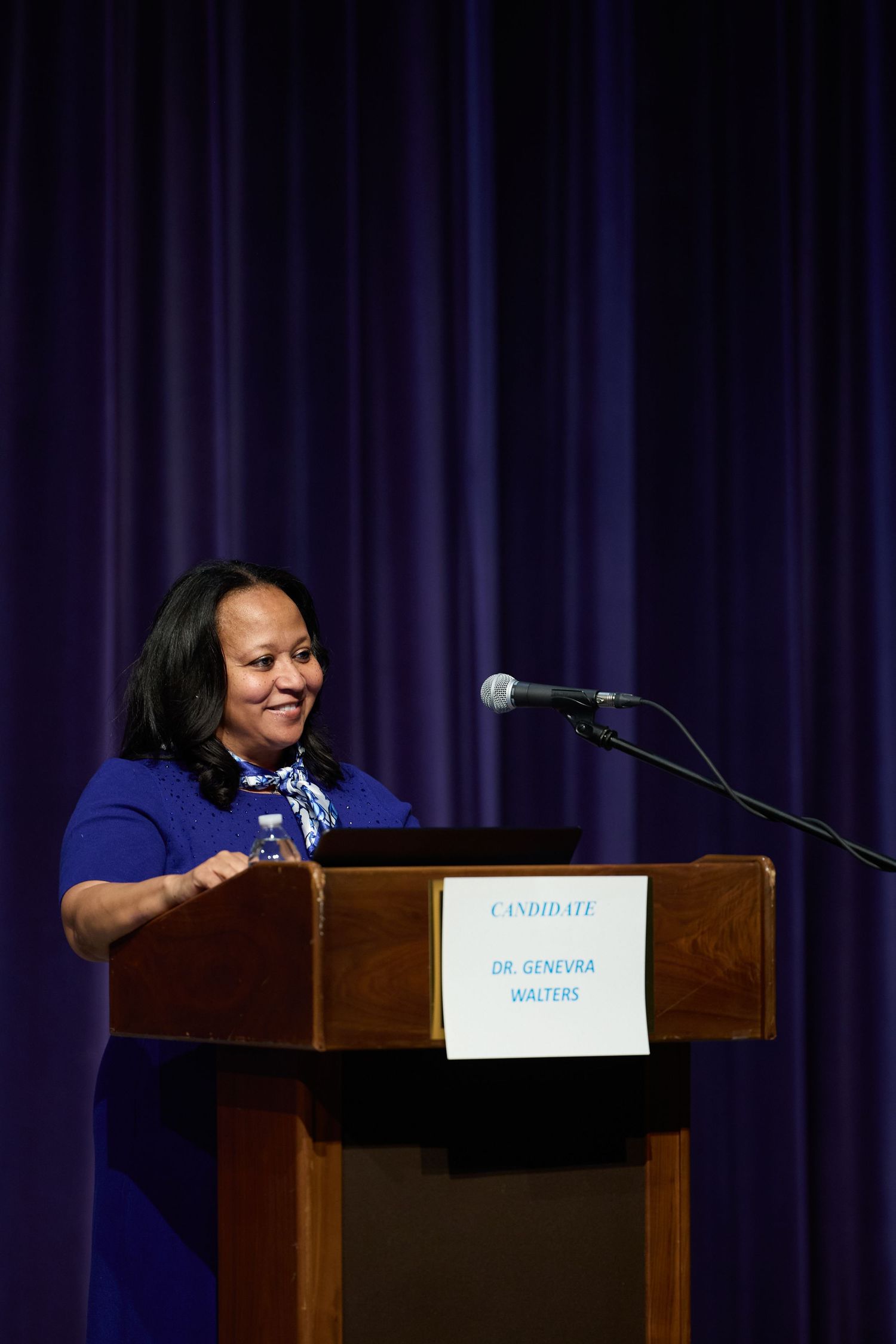 A speaker at a podium leans over and laughs during a presentation on stage with dark blue curtains behind them.