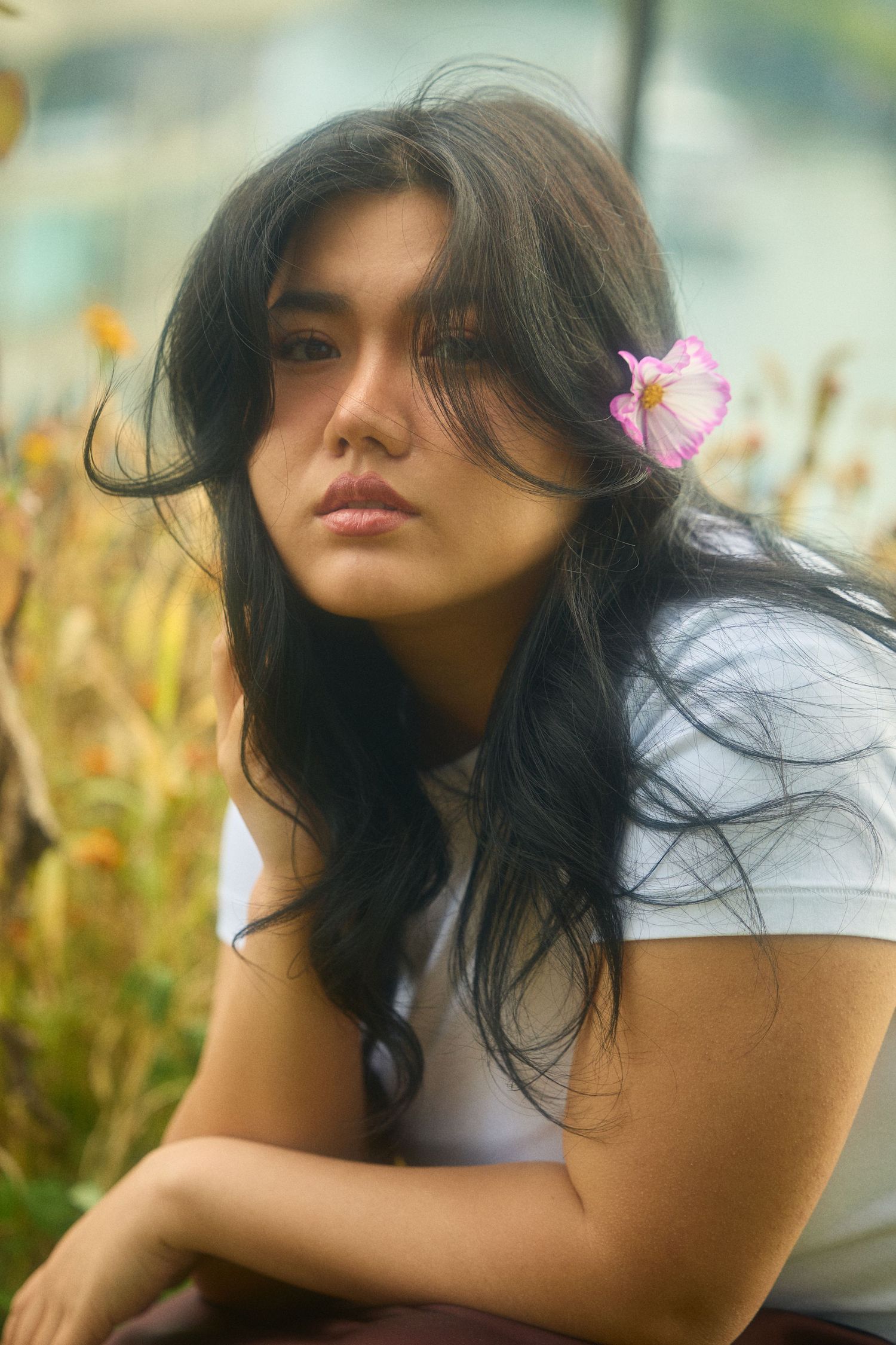 Woman with flowing dark hair poses in white top against natural outdoor backdrop with flowers.