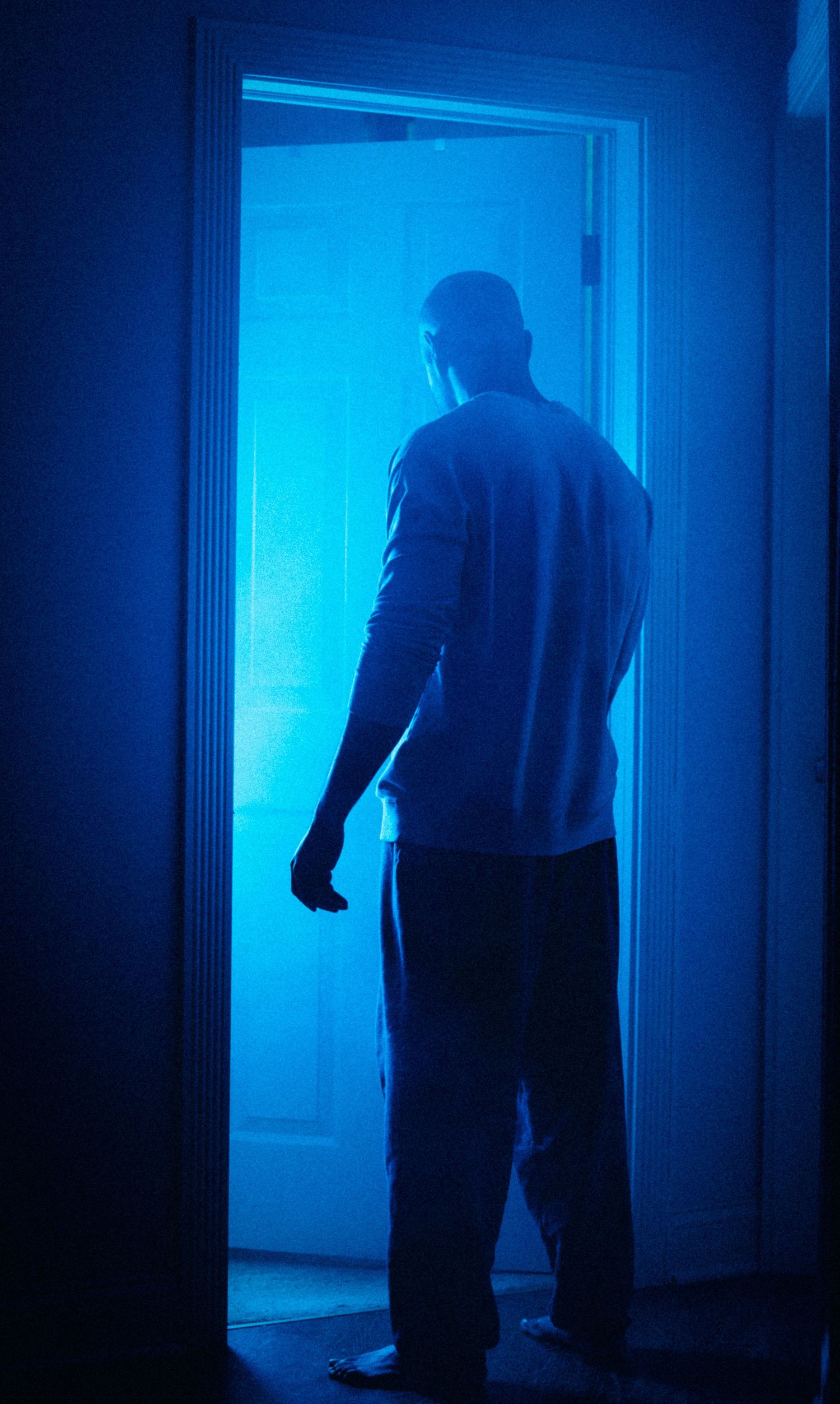A silhouetted figure stands in a doorway bathed in eerie blue light creating a mysterious atmospheric scene.