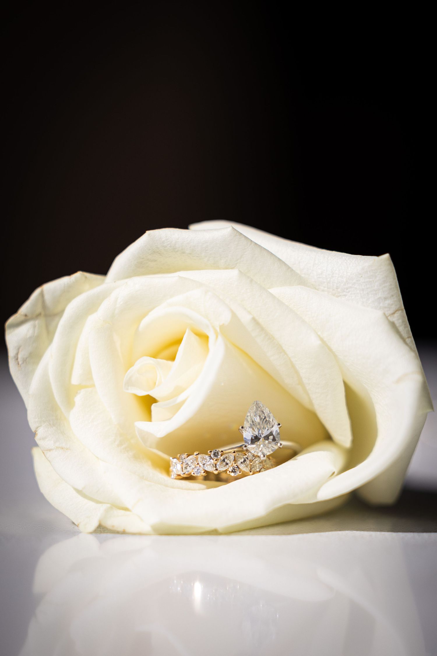 A sparkling diamond engagement ring rests elegantly on a pure white rose in a romantic close-up photograph.